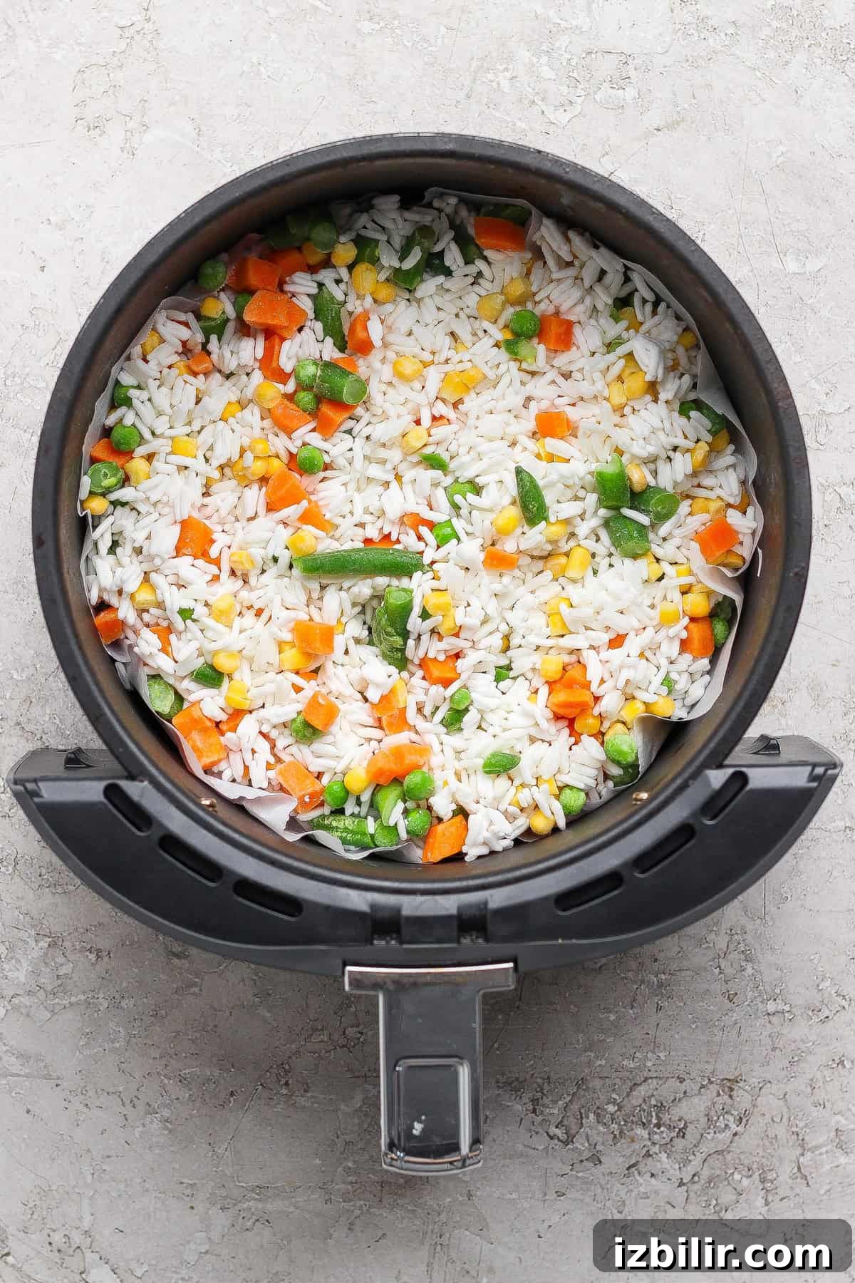An air fryer basket filled with uncooked white rice and mixed vegetables including carrots, peas, corn, and green beans.
