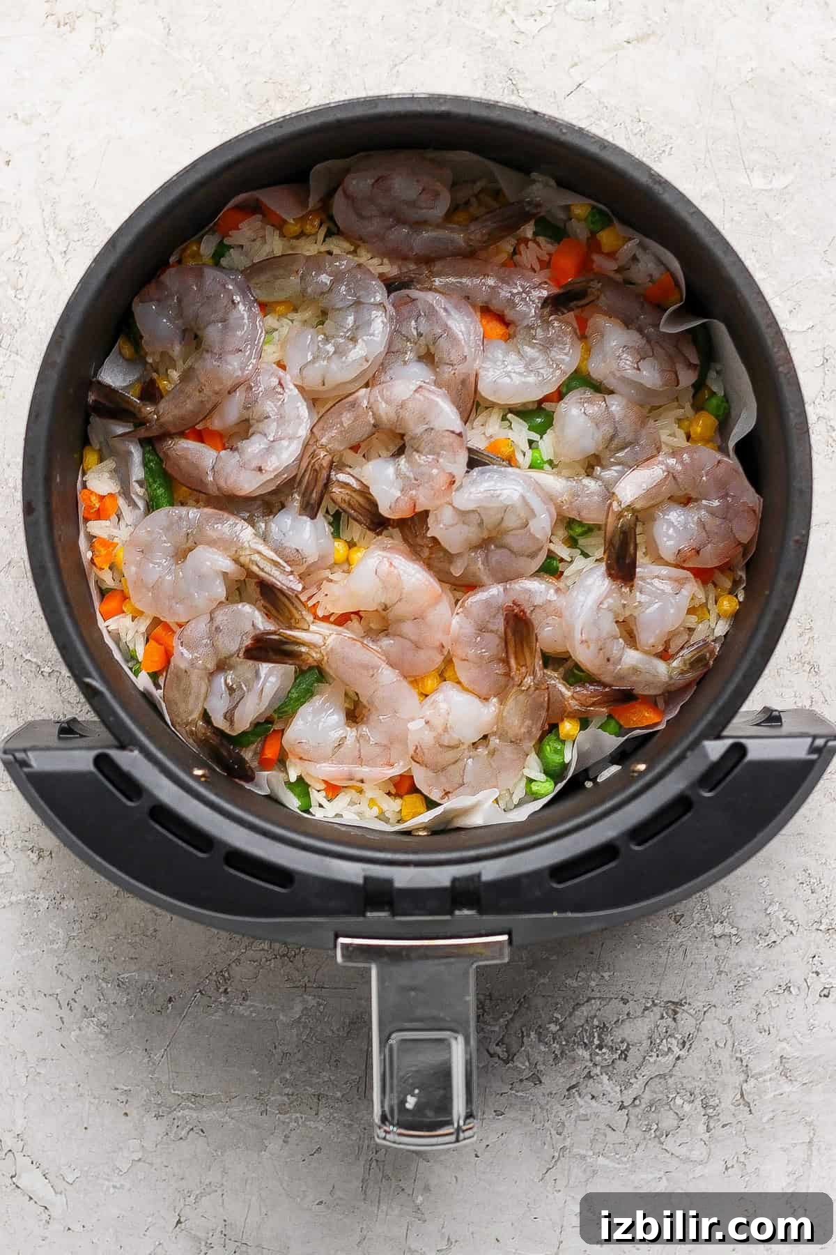 Raw shrimp arranged on top of mixed vegetables and rice in the basket of an air fryer.