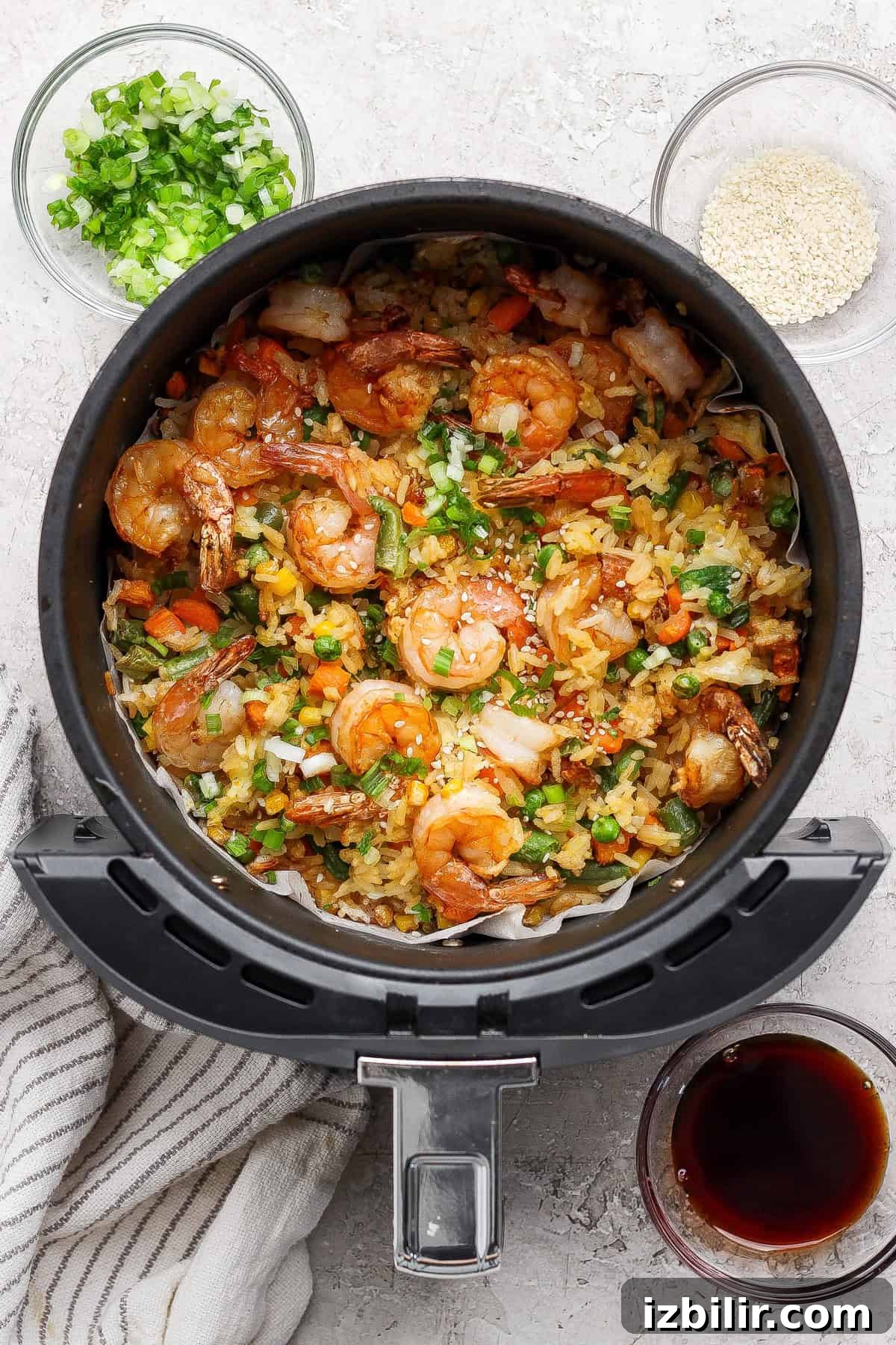 Cooked shrimp fried rice in an air fryer basket, garnished with chopped green onions, with bowls of sesame seeds, green onions, and a dark sauce nearby.