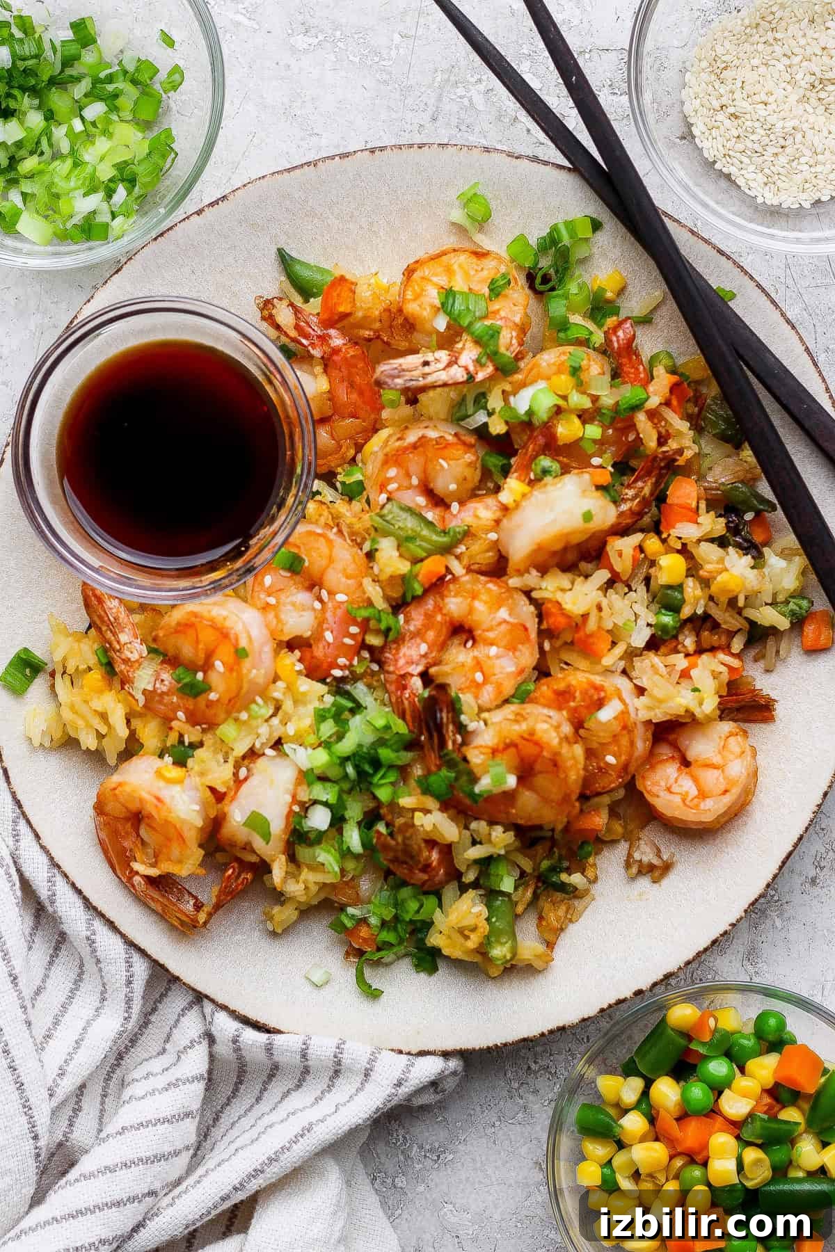 A plate of shrimp fried rice with mixed vegetables, topped with chopped green onions, served with soy sauce and chopsticks. Small bowls of sesame seeds and vegetables are beside the plate.
