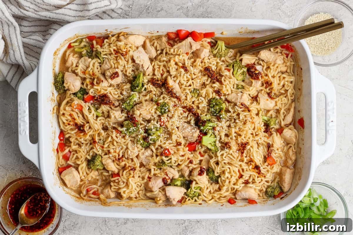 Crispy Oven-Baked Ramen Noodles 2 Rectangular white baking dish filled with cooked ramen noodles, chicken pieces, broccoli, and red peppers, garnished with sesame seeds and sauce, with chopsticks resting on the dish.