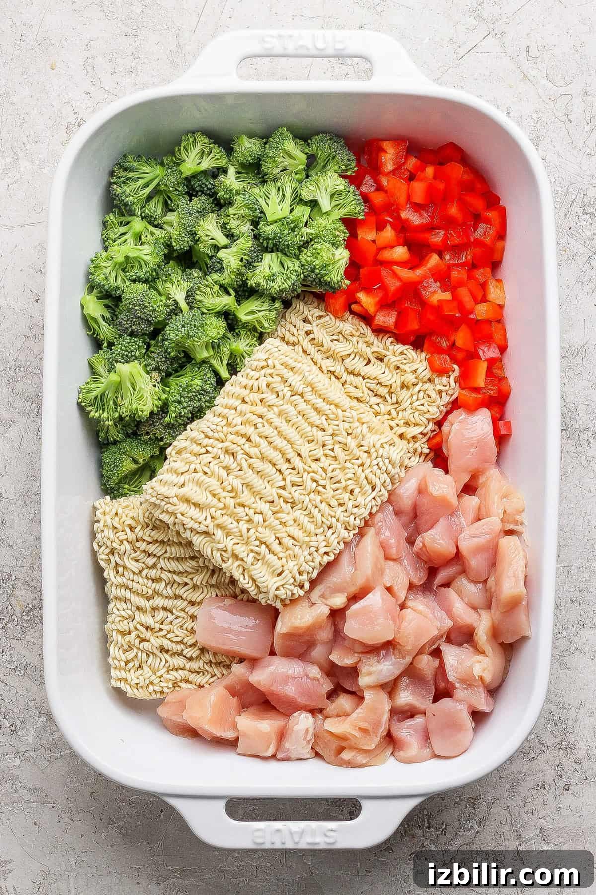 Crispy Oven-Baked Ramen Noodles 3 A white baking dish containing raw broccoli florets, diced red bell pepper, two blocks of dry ramen noodles, and chunks of raw chicken, all arranged separately.