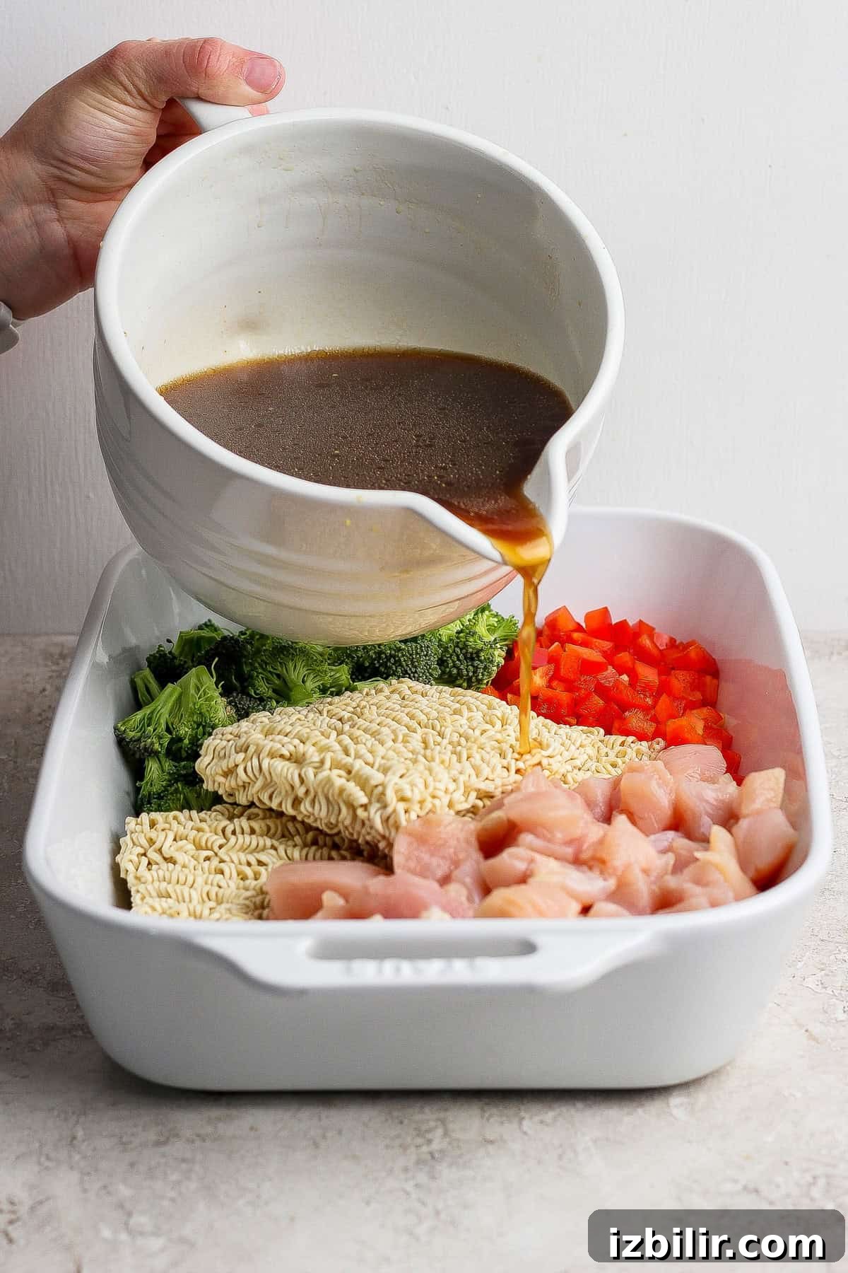 Crispy Oven-Baked Ramen Noodles 4 A hand pours brown sauce from a white bowl over uncooked ramen noodles, chopped broccoli, red bell pepper, and diced chicken in a white baking dish.