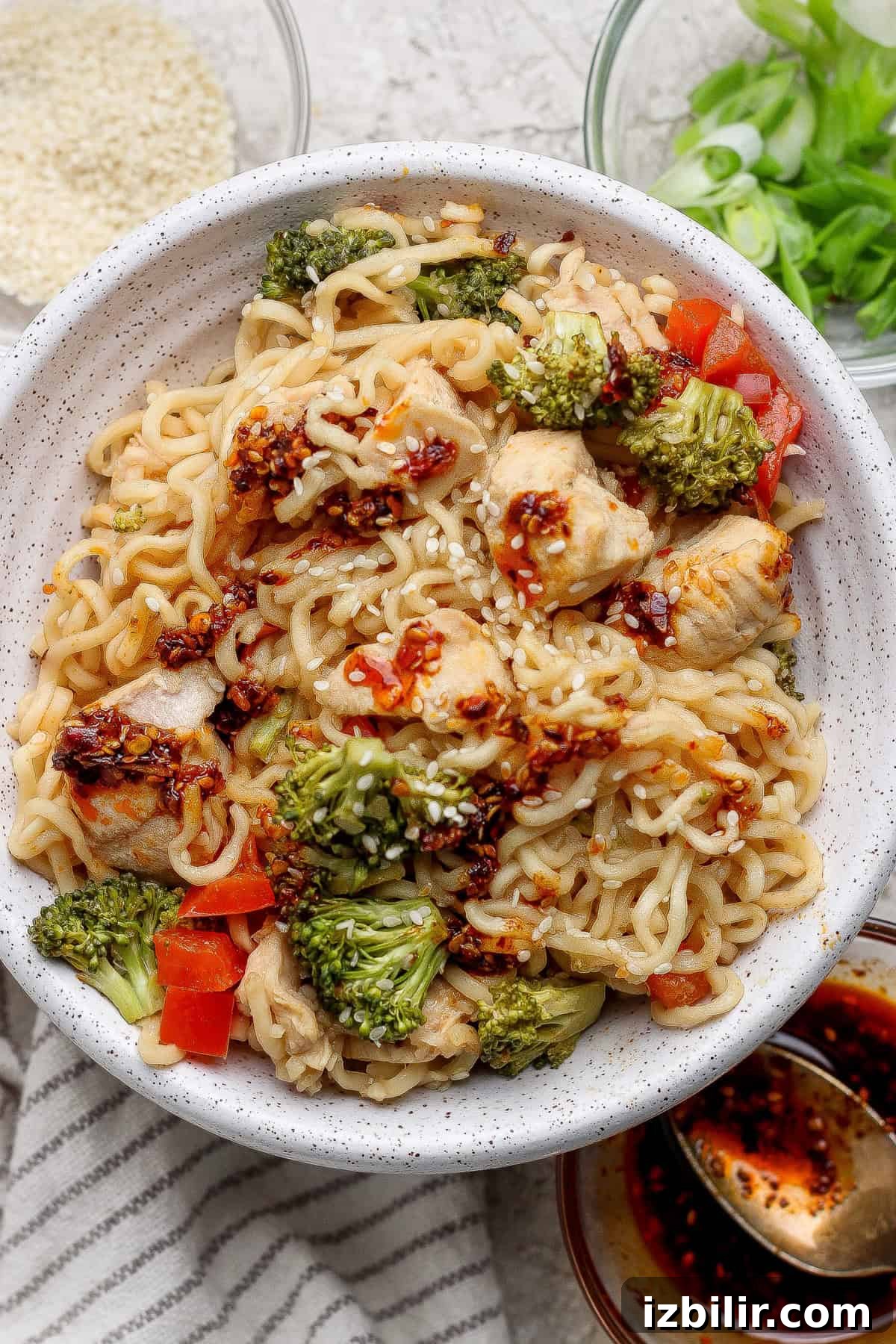 Crispy Oven-Baked Ramen Noodles 5 A bowl of ramen noodles mixed with chicken, broccoli, and red bell peppers, topped with chili oil and sesame seeds, with green onions and sauces on the side.