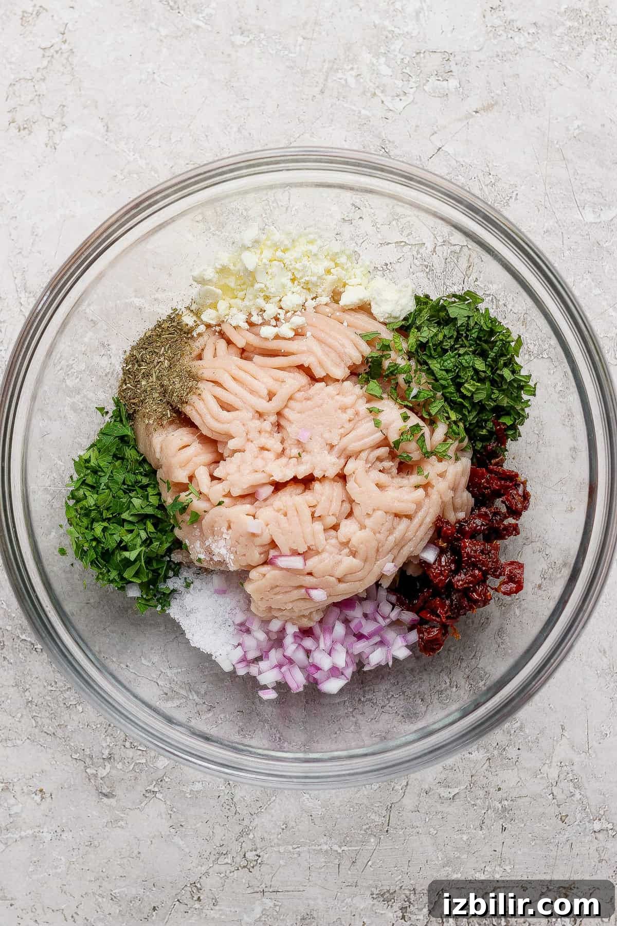 A glass bowl containing ground meat, chopped red onion, herbs, minced garlic, sun-dried tomatoes, salt, pepper, and seasonings arranged separately.
