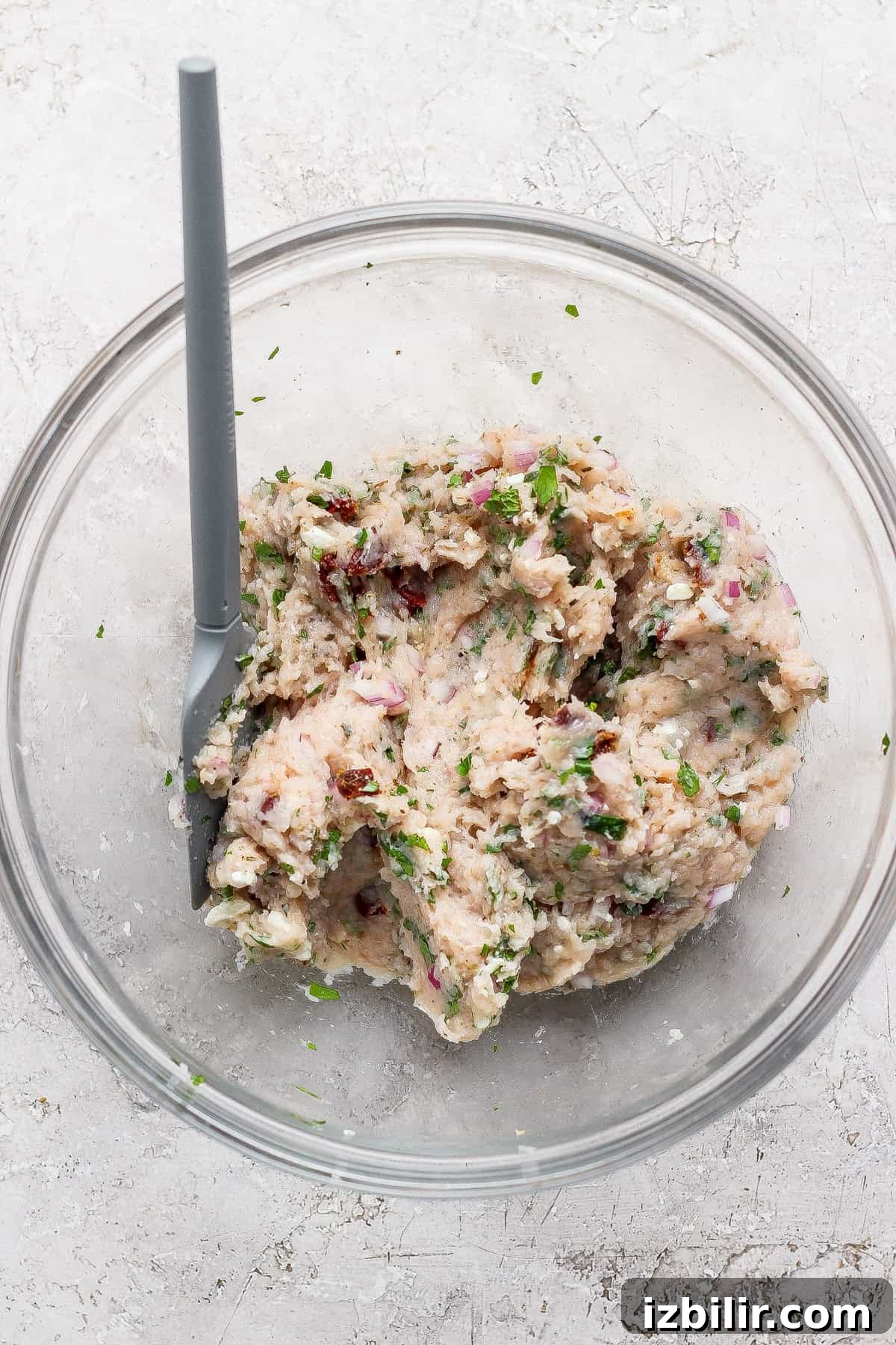 A glass bowl with a gray spoon contains a mixture of mashed white beans, chopped herbs, red onion, and sun-dried tomatoes on a light-colored surface.