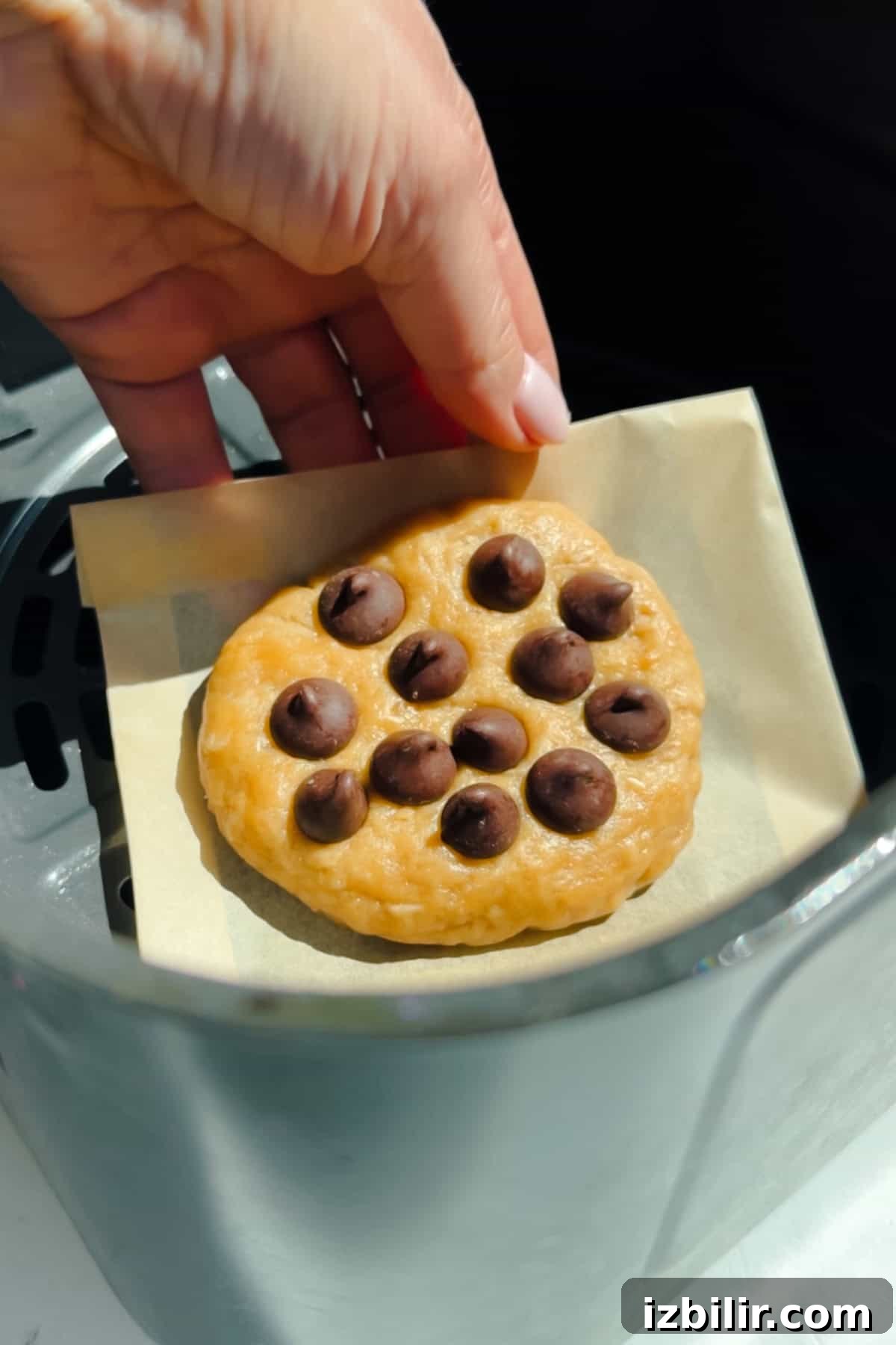 A hand is placing a round chocolate chip cookie dough on a piece of parchment paper inside an air fryer basket.