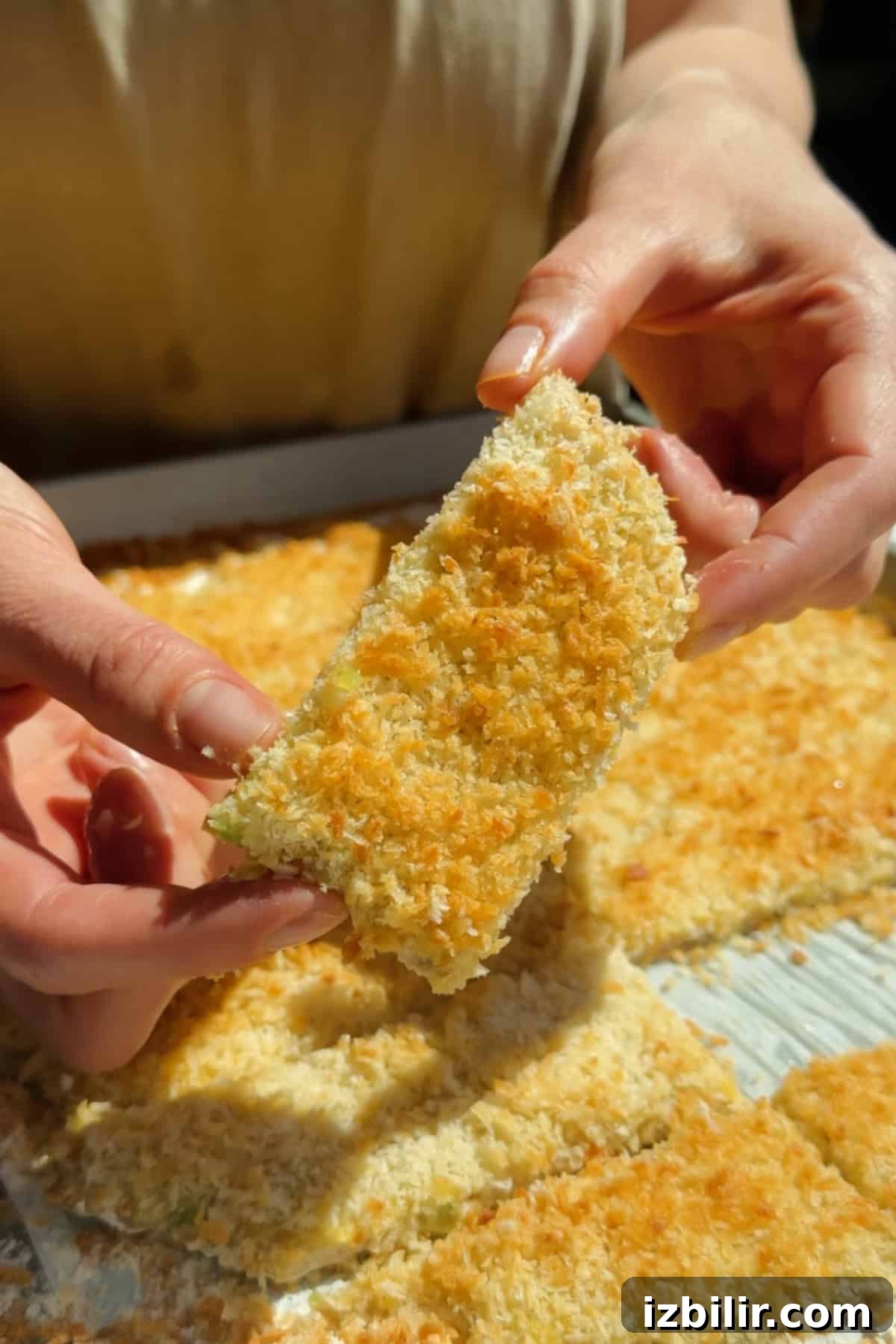 Holding a golden crispy chicken protein bar with panko coating above a tray of baked bars