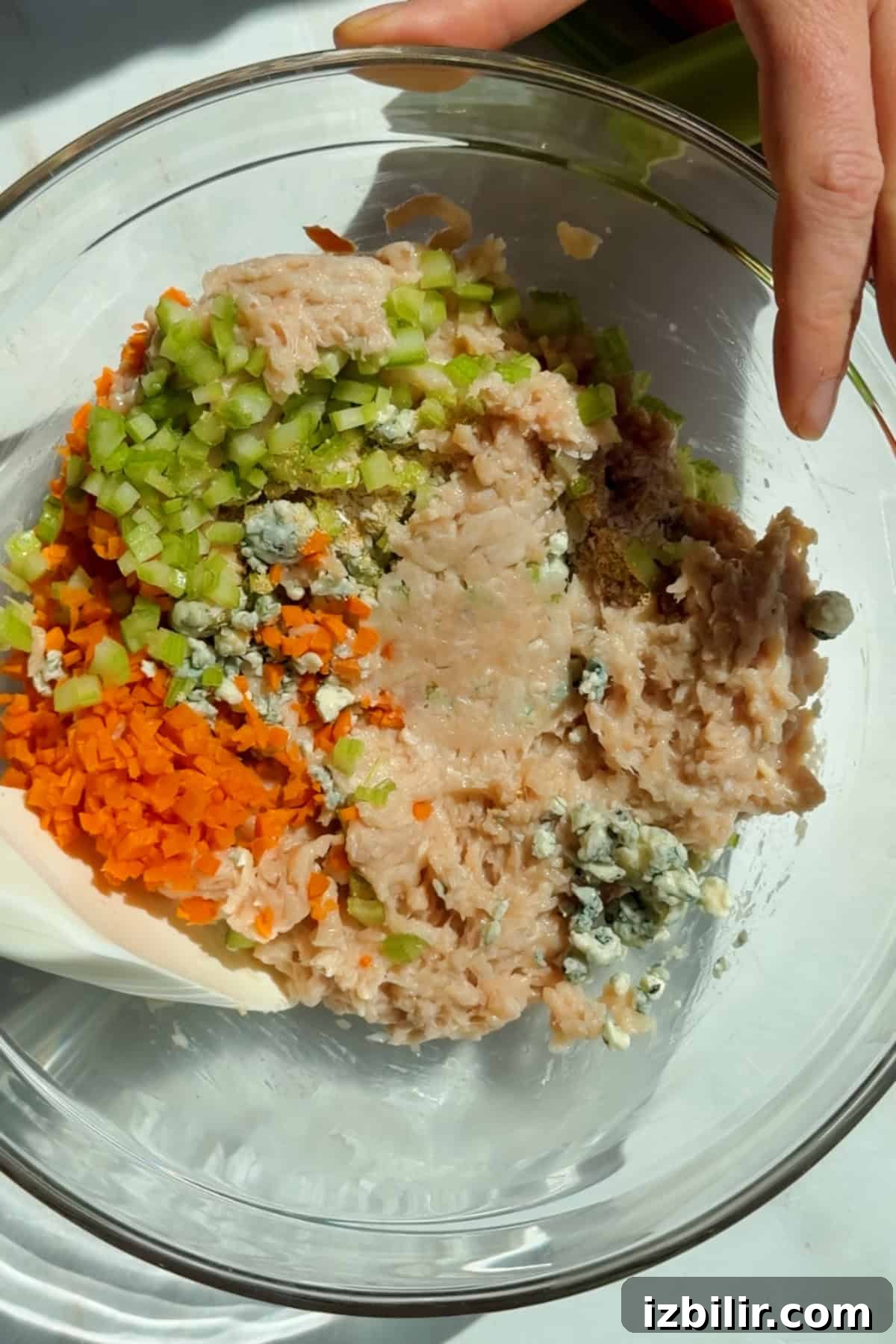 Mixing ground chicken, carrots, celery, blue cheese crumbles, and seasonings in a glass bowl for protein bars