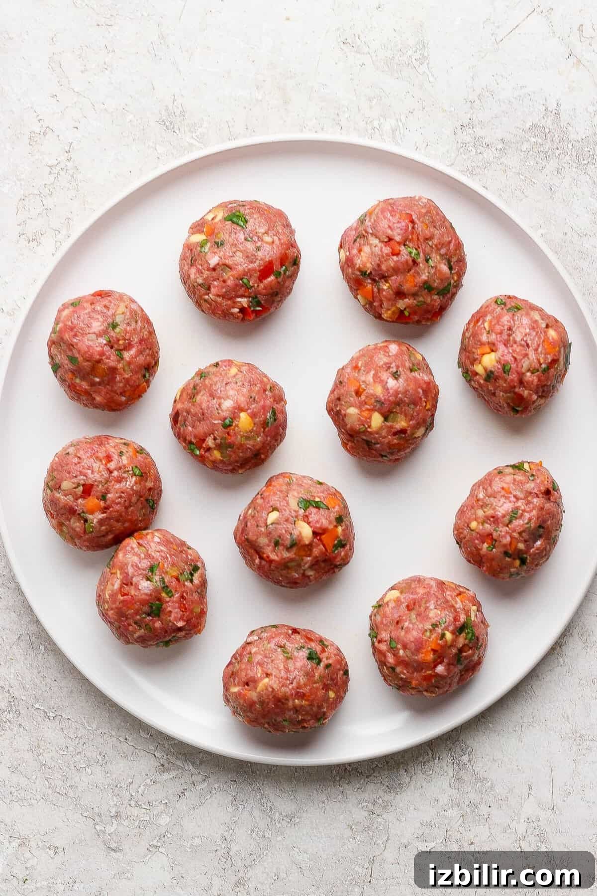 A white plate with twelve uncooked meatballs, mixed with herbs and small vegetable pieces, on a light textured surface.