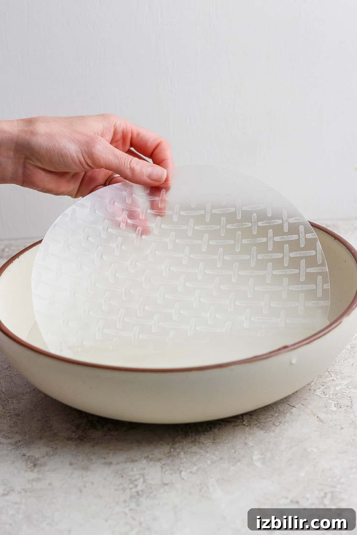 A hand dips a round sheet of rice paper into a shallow bowl of water on a light-colored surface.