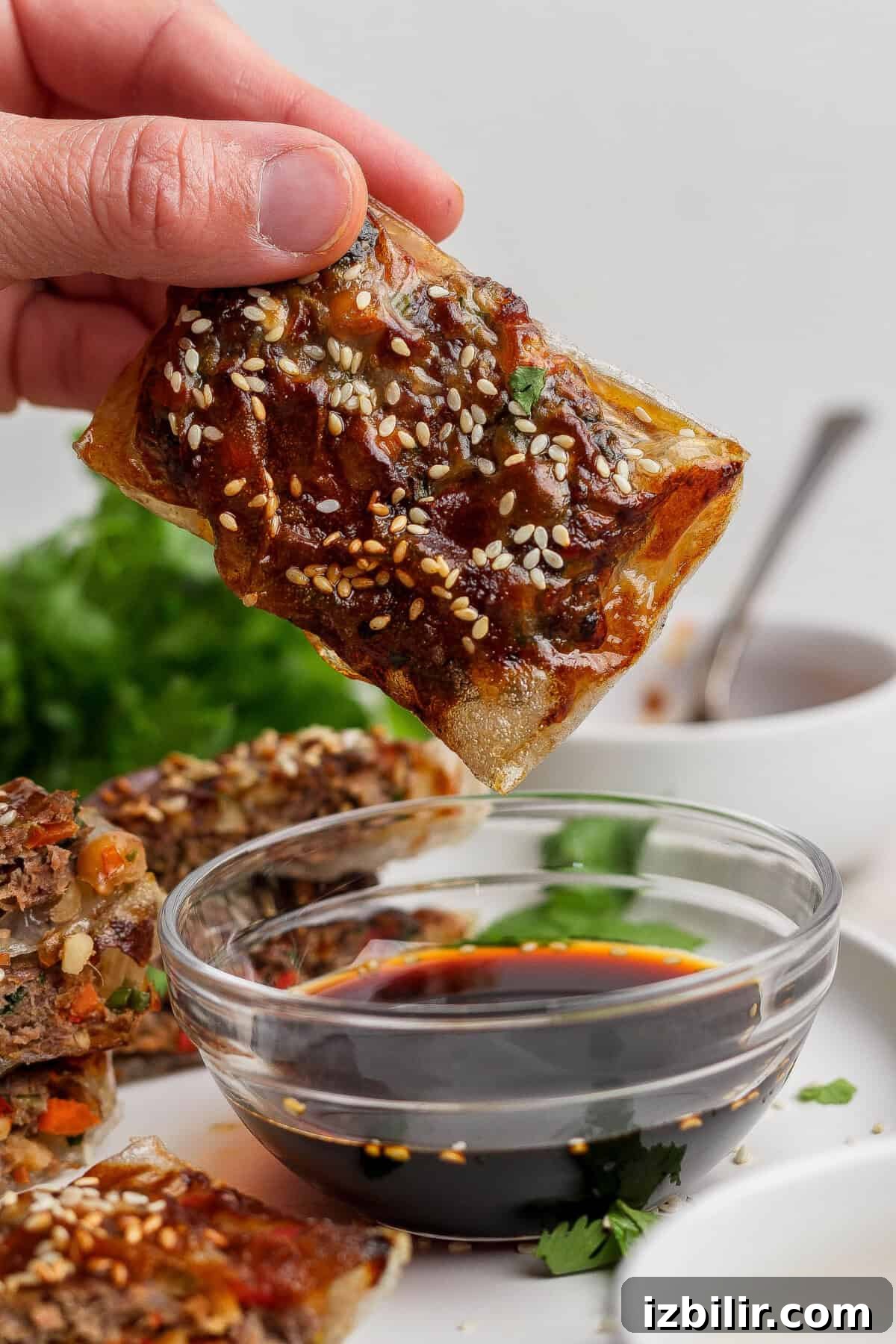 A hand holds a crispy, rectangular pastry topped with sesame seeds, about to dip it into a bowl of soy sauce; greens and more pastries are in the background.