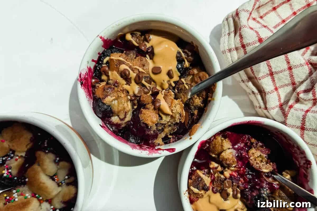 2-Ingredient Air Fryer Berry Cobbler 2 Three small white ramekins filled with baked berry dessert, topped with peanut butter and chocolate chips, with metal spoons and a red-striped cloth nearby.