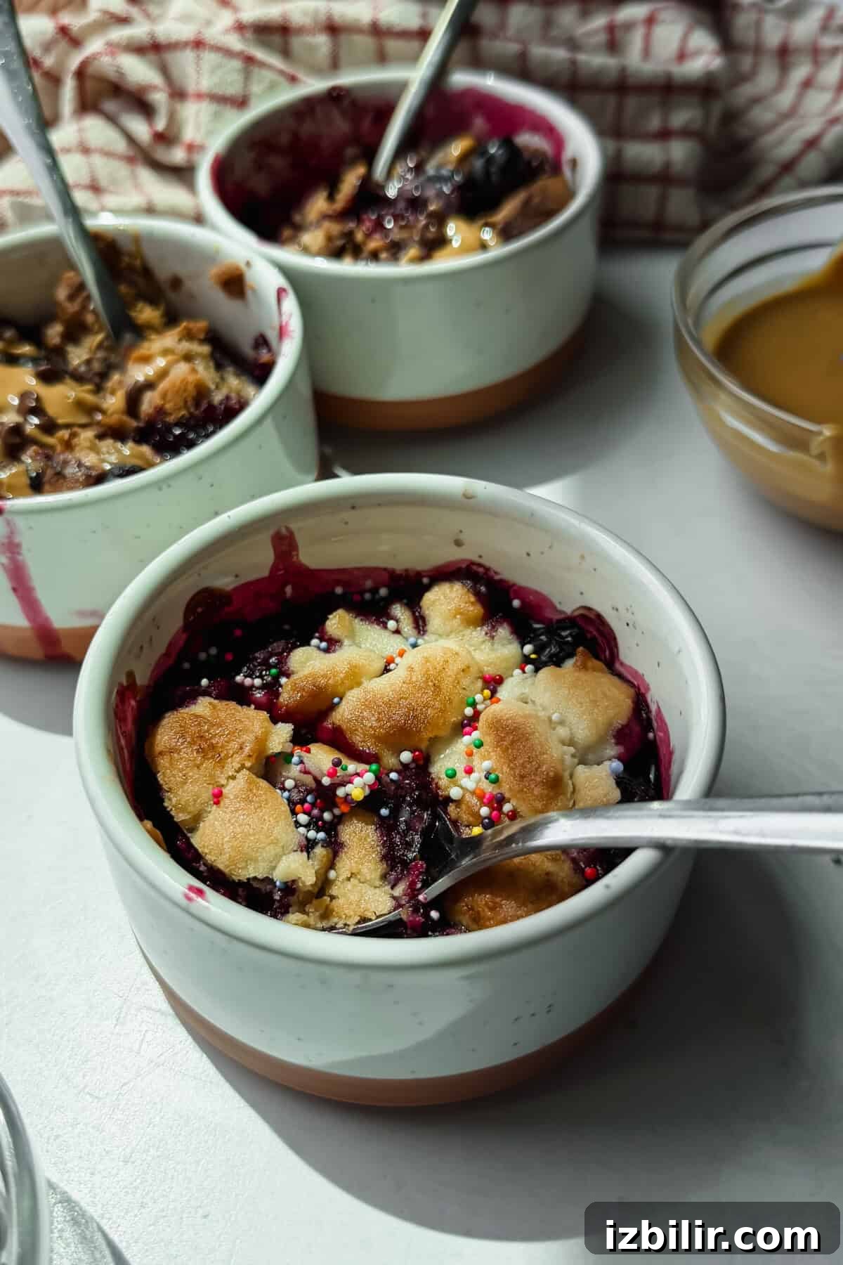 2-Ingredient Air Fryer Berry Cobbler 3 Three ceramic bowls of berry crumble topped with sprinkles and shortbread pieces, each with a spoon, sit on a light surface next to a bowl of caramel sauce.