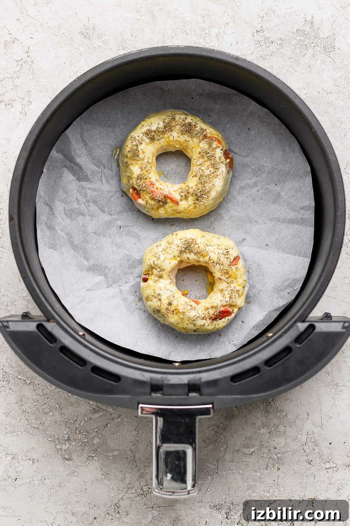 Two uncooked pizza protein bagels with seasoning in an air fryer basket