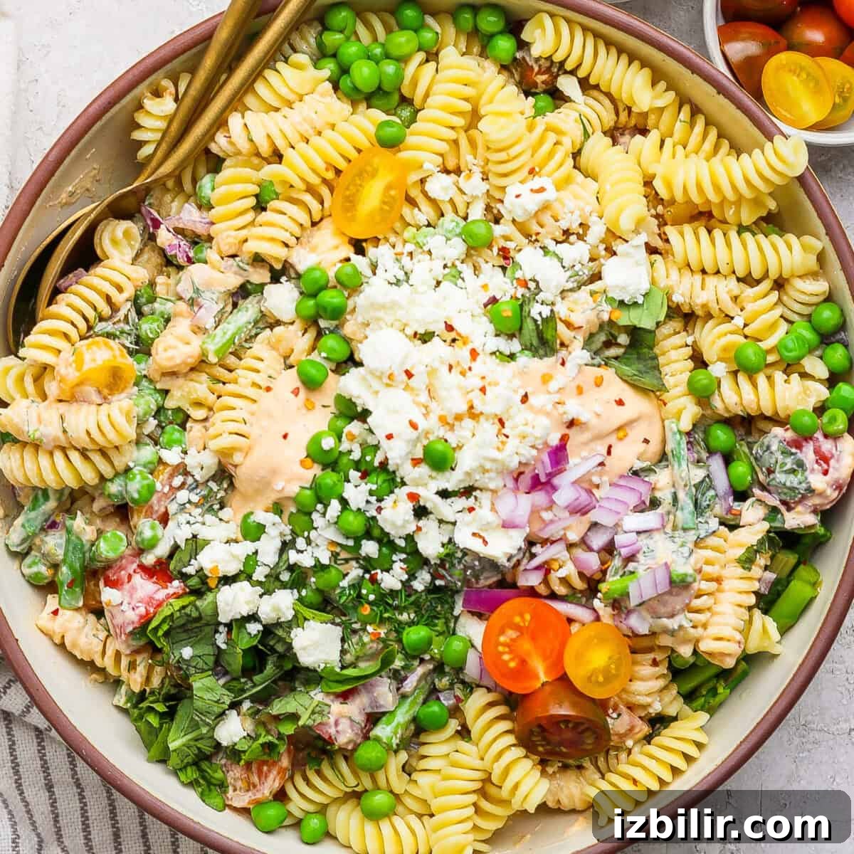 Bowl of pasta salad with rotini, peas, cherry tomatoes, crumbled cheese, greens, and a creamy dressing, garnished with chopped onions and red pepper flakes.
