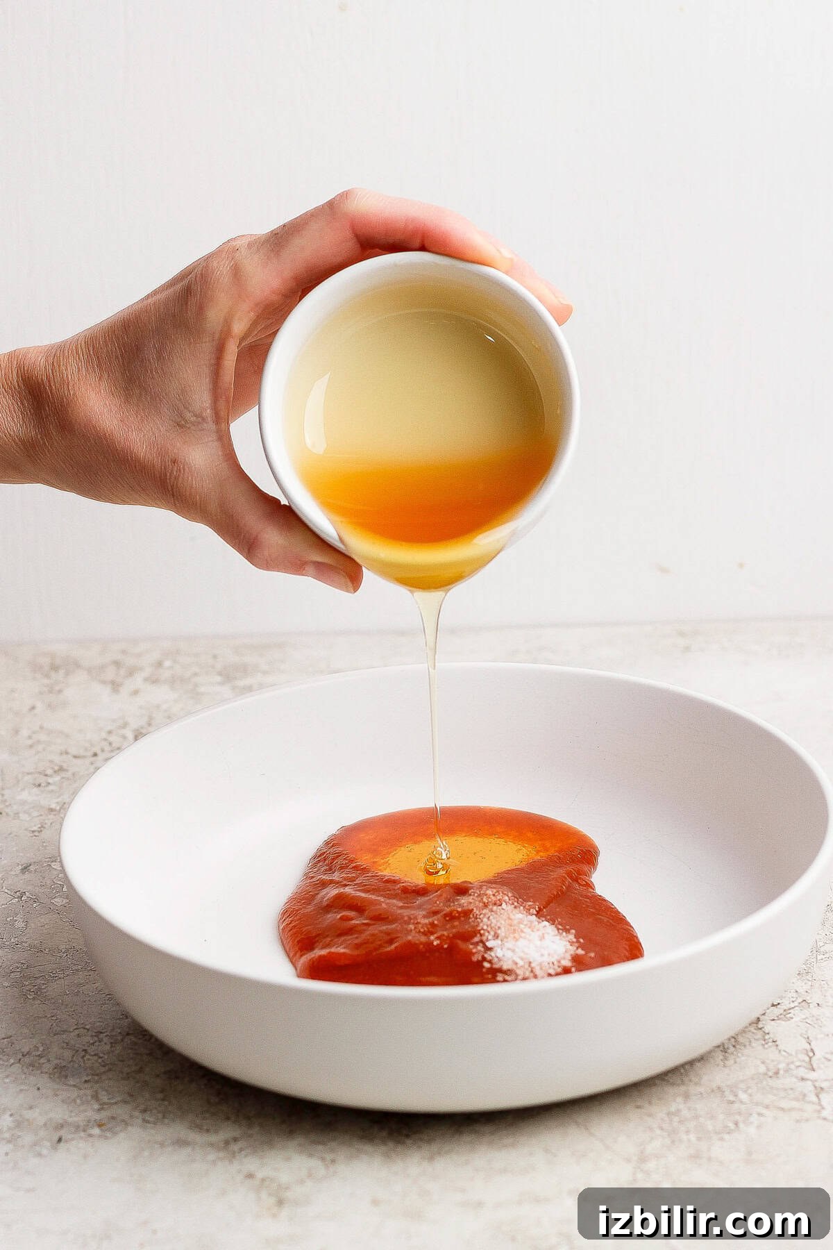 A hand pours honey from a small white bowl onto a plate of barbecue sauce.