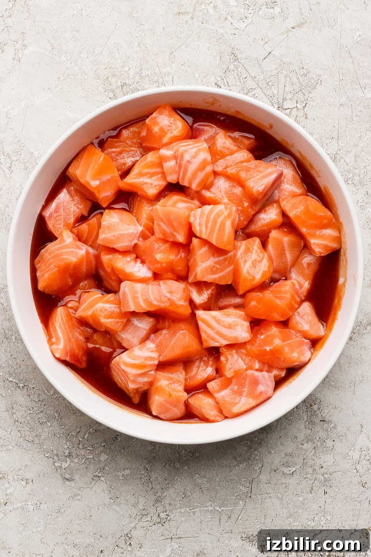 A white bowl filled with cubed raw salmon mixed with a red marinade, placed on a light textured surface.