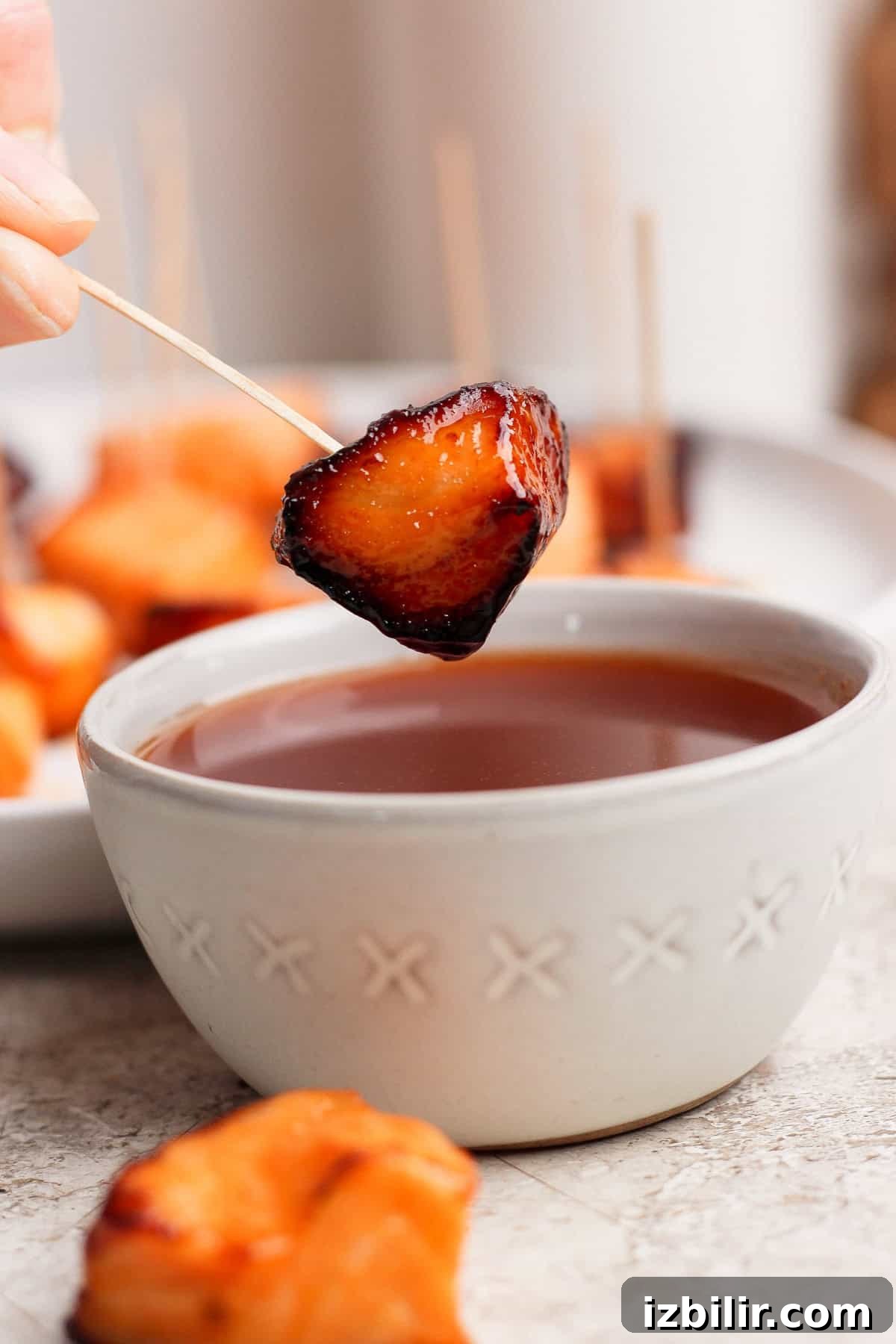 A hand holds a skewer with a glazed, roasted bite-sized food piece above a bowl of dipping sauce; more skewered pieces are seen in the background.