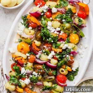 A white platter with gnocchi, roasted cherry tomatoes, bell peppers, red onions, fresh greens, and crumbled feta cheese.