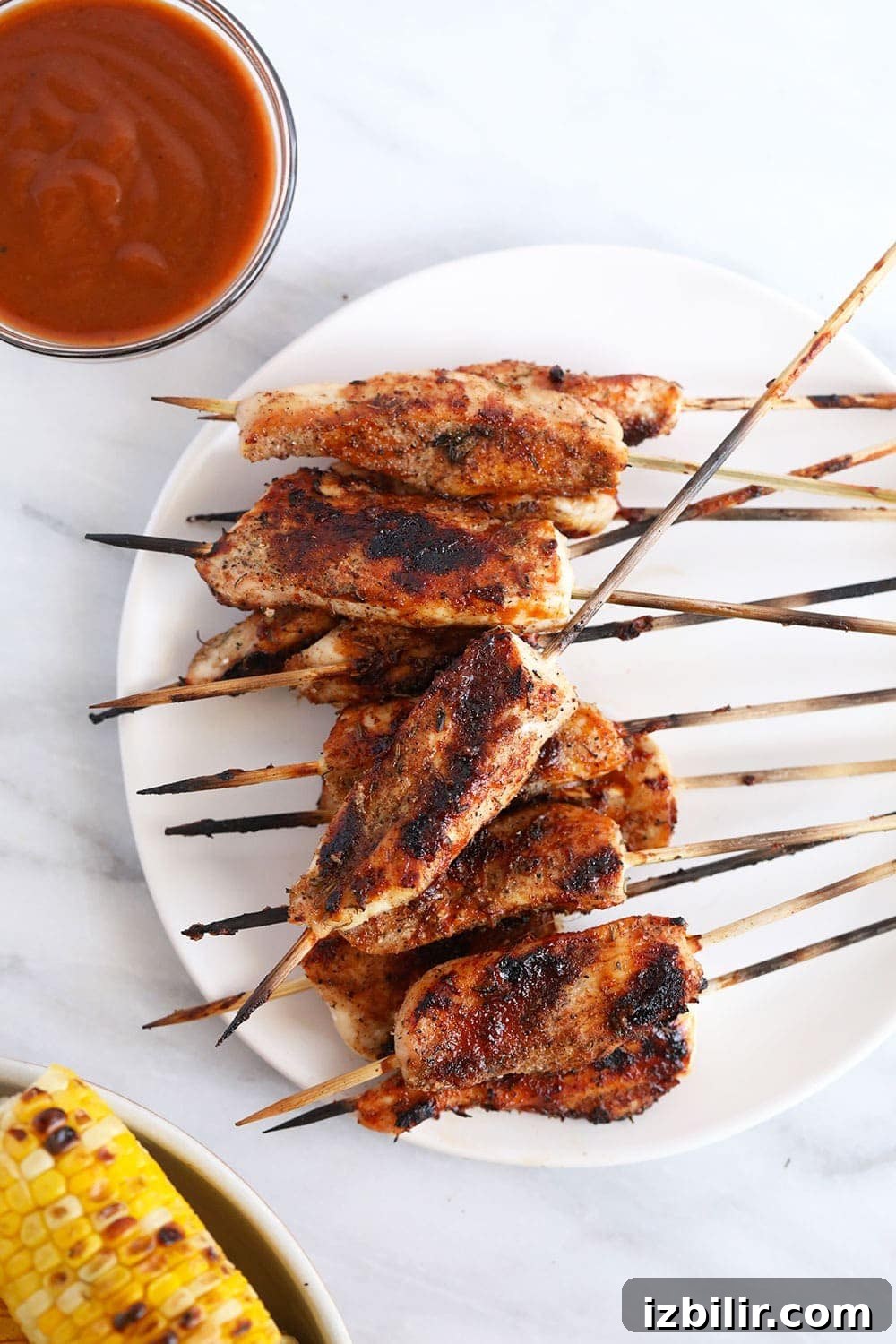These BBQ Chicken Skewers are the perfect summer meal! Grab some skewers, BBQ sauce, and your favorite BBQ side dish and let's grill! Grilled skewers on plate next to bbq sauce and grilled corn.