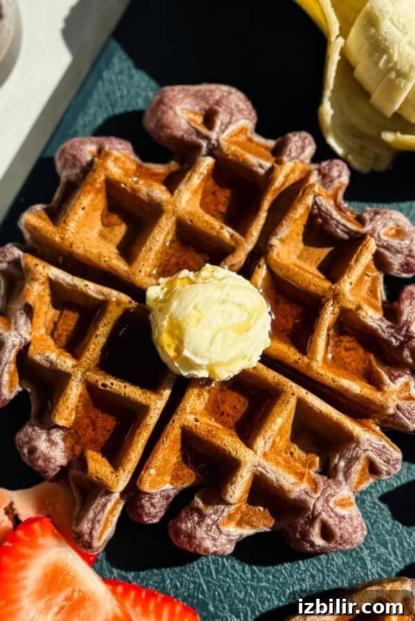 A waffle topped with a dollop of butter is surrounded by sliced bananas and strawberries on a dark surface.