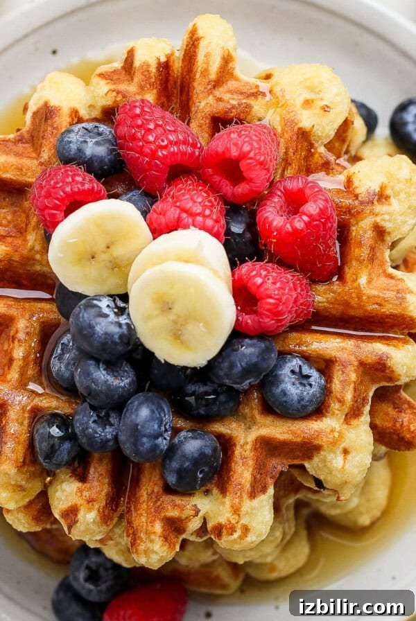 A stack of waffles topped with blueberries, raspberries, banana slices, and drizzled with syrup on a round white plate.