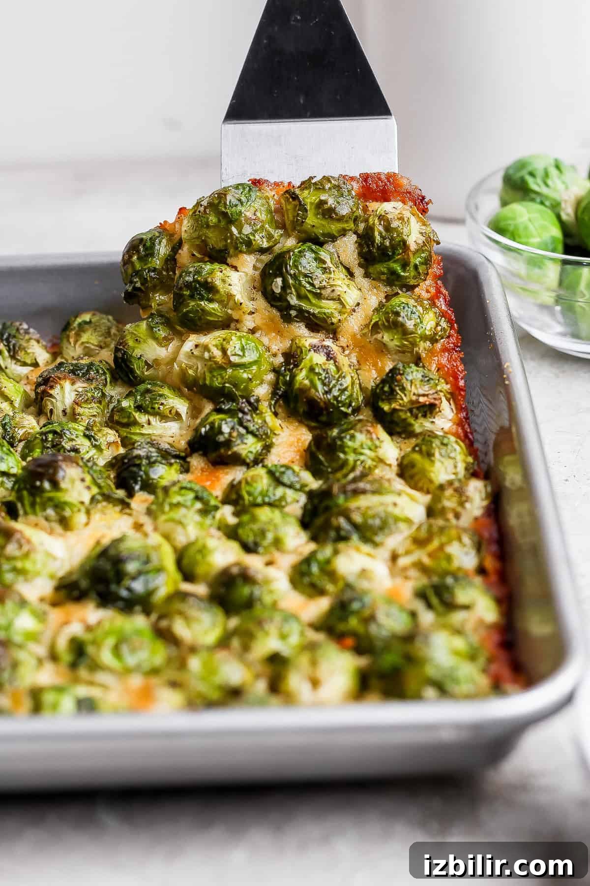 Crispy Parmesan-Crusted Brussels Sprouts Recipe 7 A metal spatula lifts a piece of baked casserole topped with roasted brussels sprouts from a rectangular baking pan. A bowl of raw brussels sprouts sits in the background.