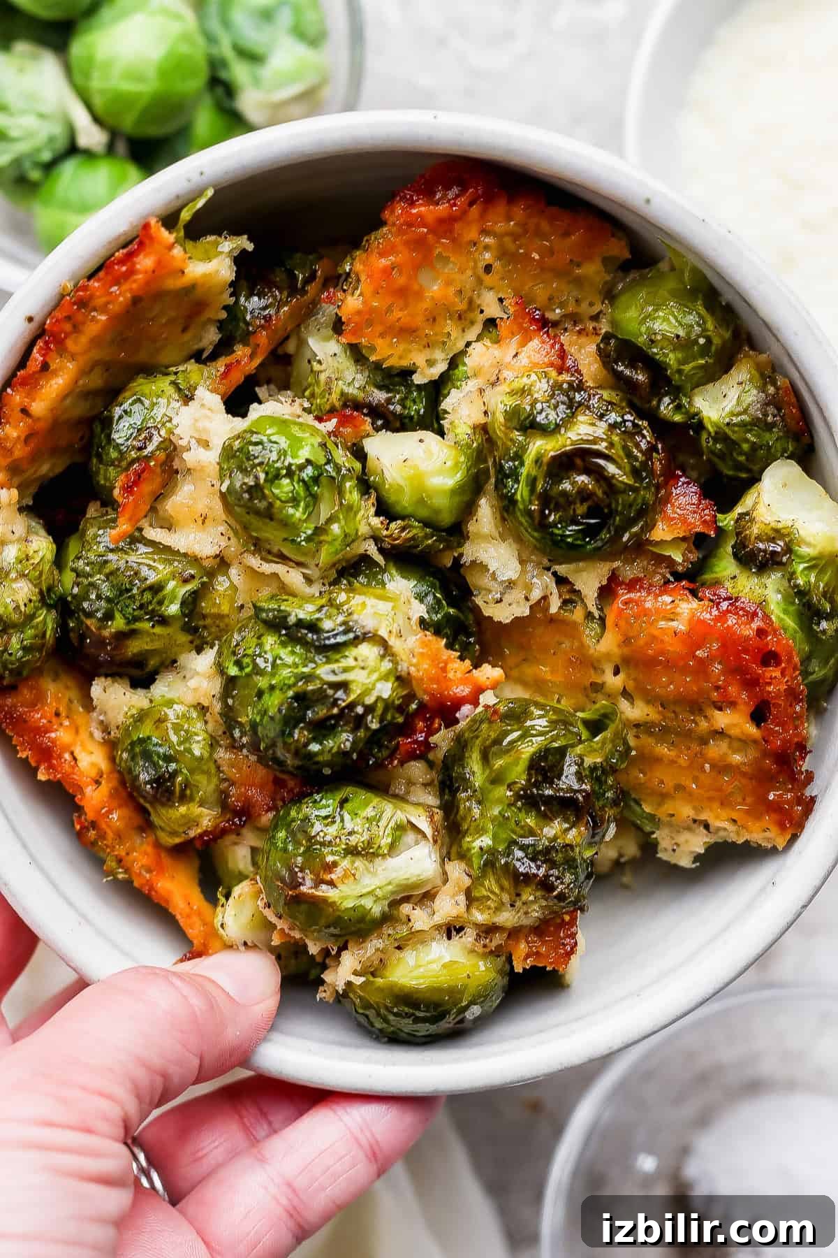 Crispy Parmesan-Crusted Brussels Sprouts Recipe 8 A bowl of roasted Brussels sprouts topped with crispy, golden-brown cheese, with a hand holding the bowl.