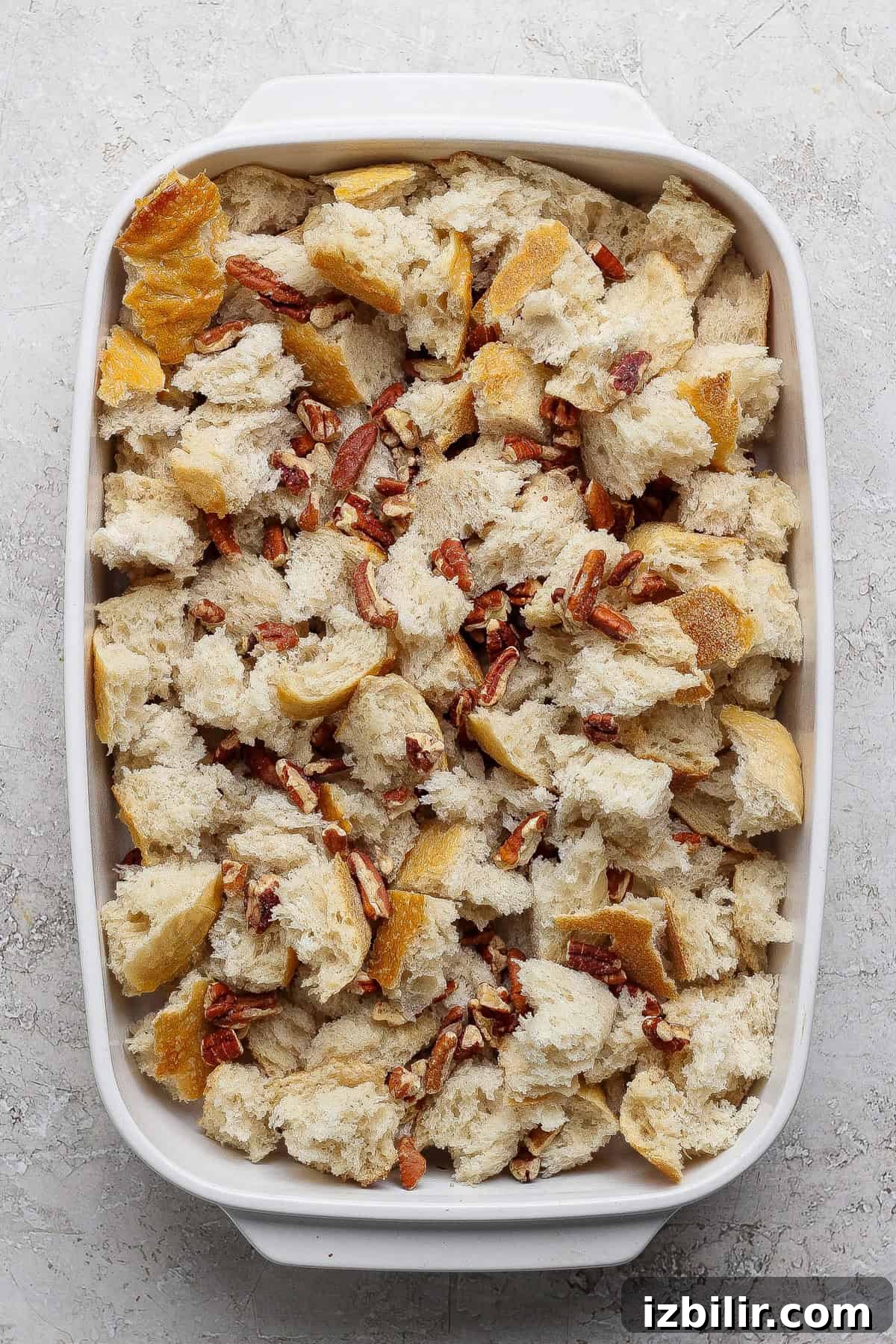 A white baking dish filled with chunks of bread and chopped pecans on a light-colored surface.