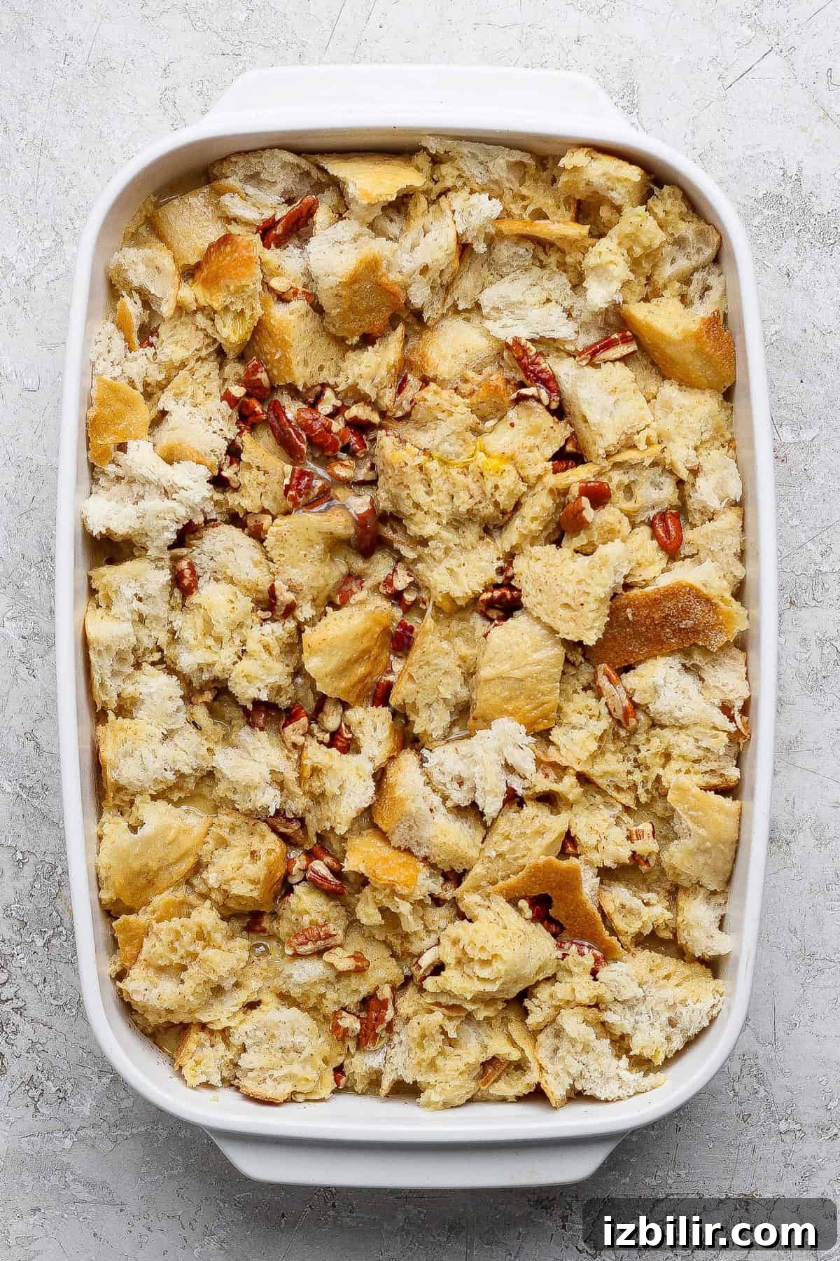 A white baking dish filled with unbaked bread pudding made of torn bread pieces and pecans on a light textured surface.
