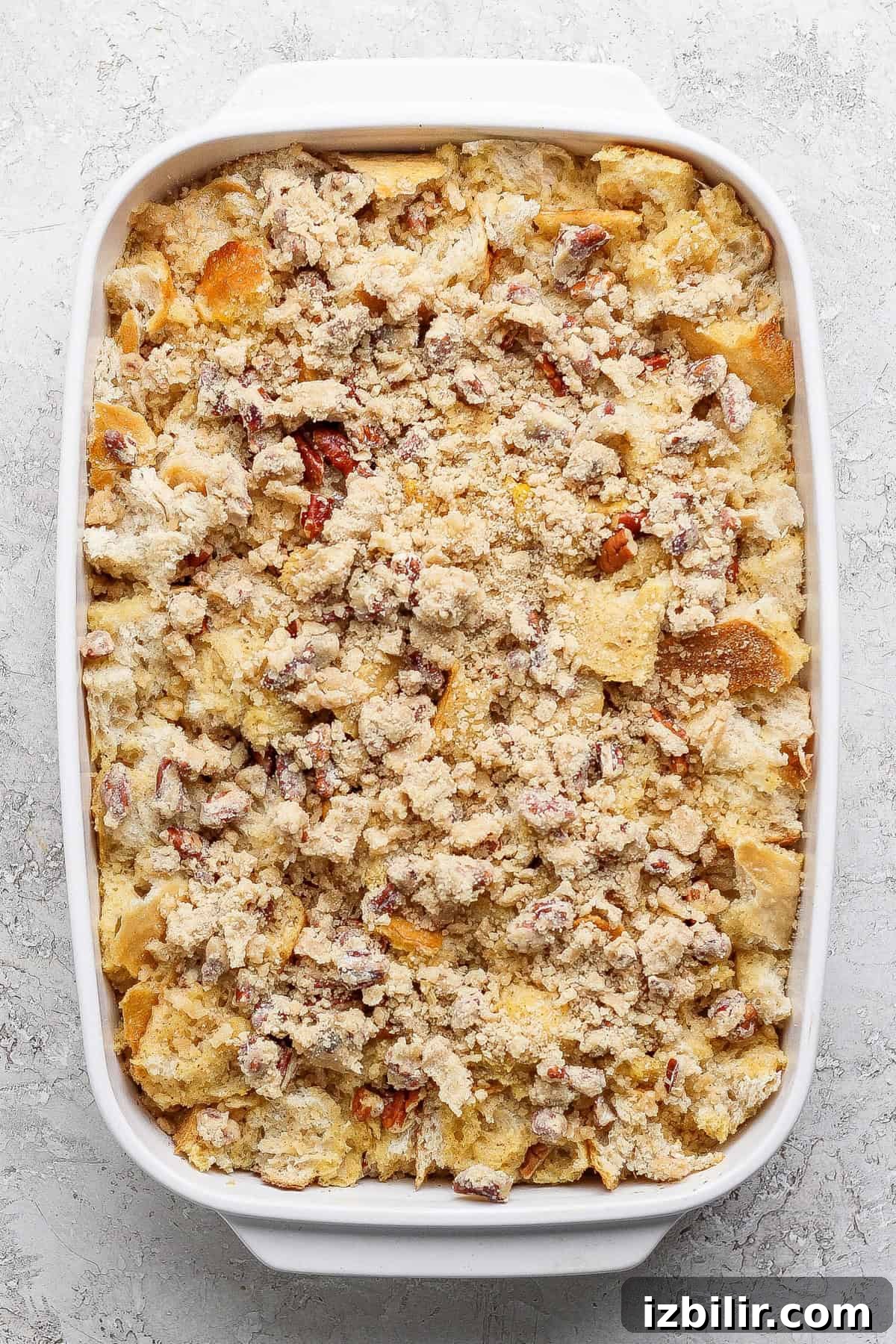 A rectangular white baking dish filled with a crumbly casserole or bread pudding mixture, topped with chopped nuts or streusel, on a light textured surface.