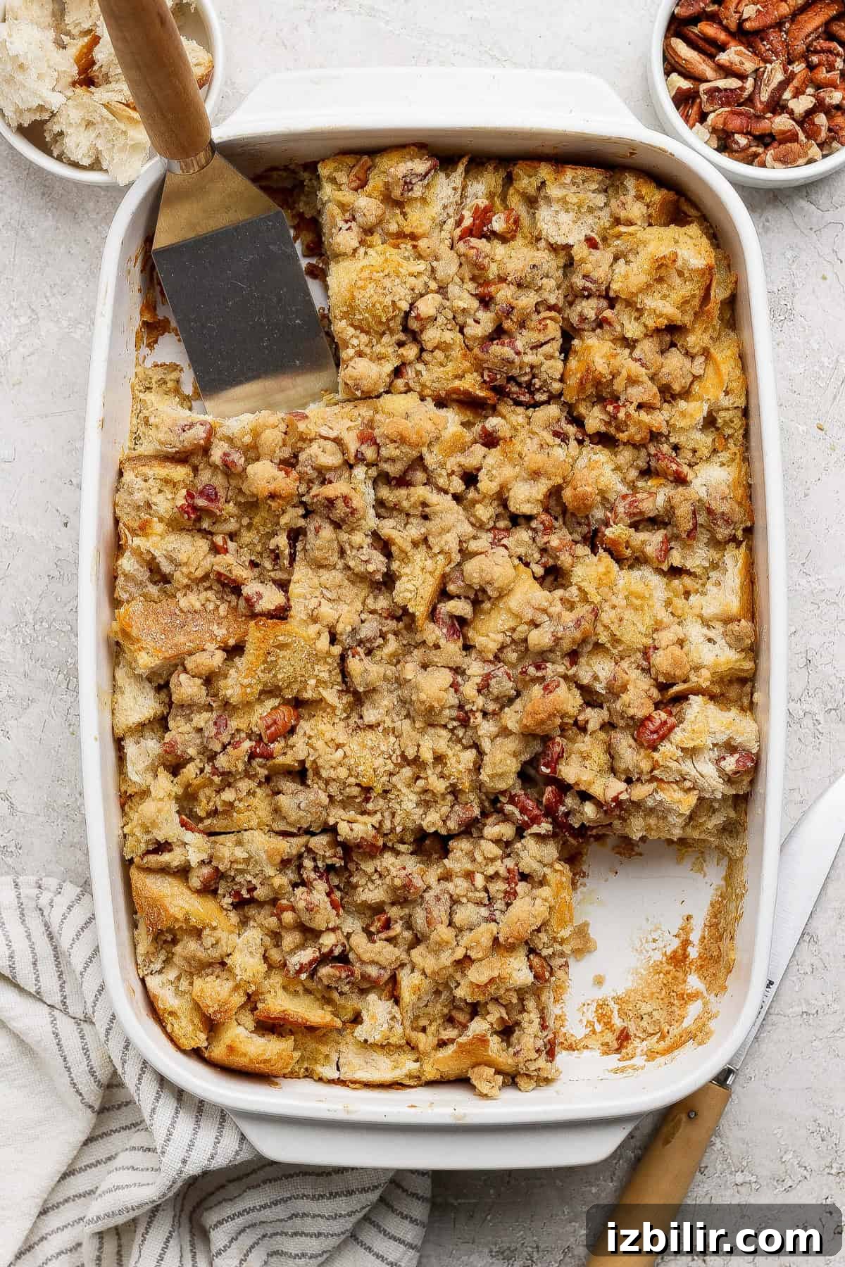 A baked casserole dish filled with pecan-topped bread pudding, partially sliced, with a spatula and a knife nearby. Small bowls of pecans and bread pieces are visible in the background.