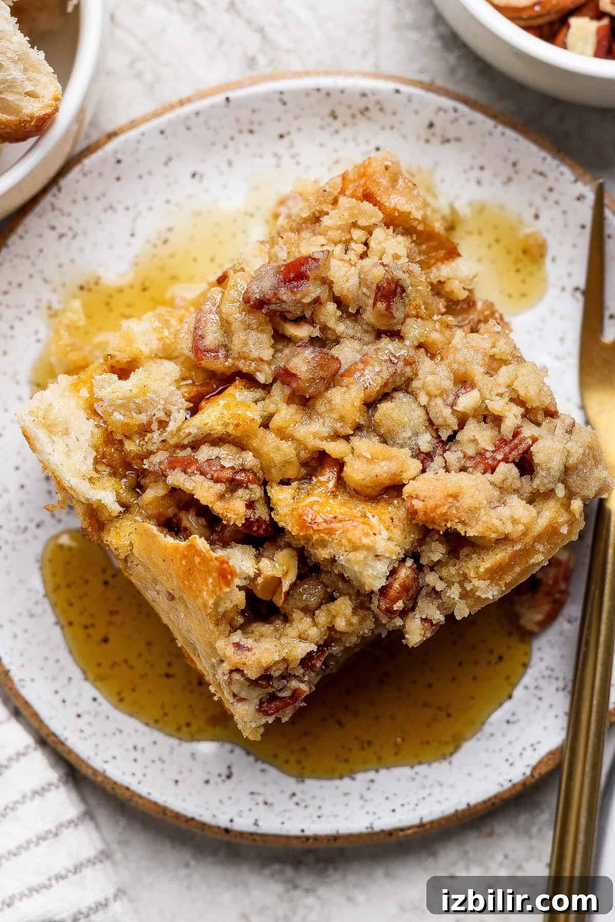 A square slice of pecan-topped bread pudding sits on a speckled plate with syrup drizzled around it and a gold fork nearby.