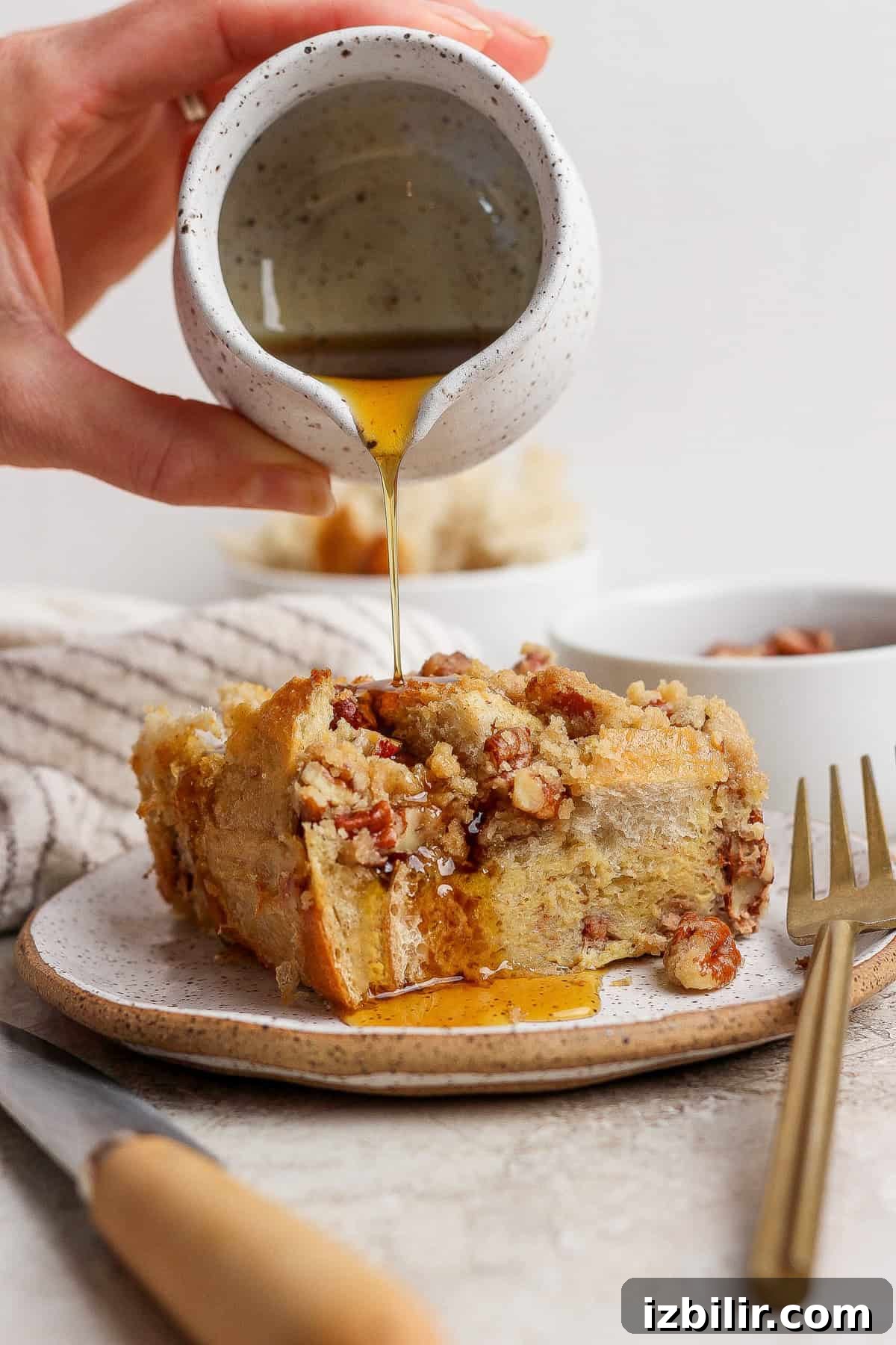 A hand pours syrup from a small pitcher onto a slice of bread pudding topped with nuts on a plate, with a fork and knife beside it.