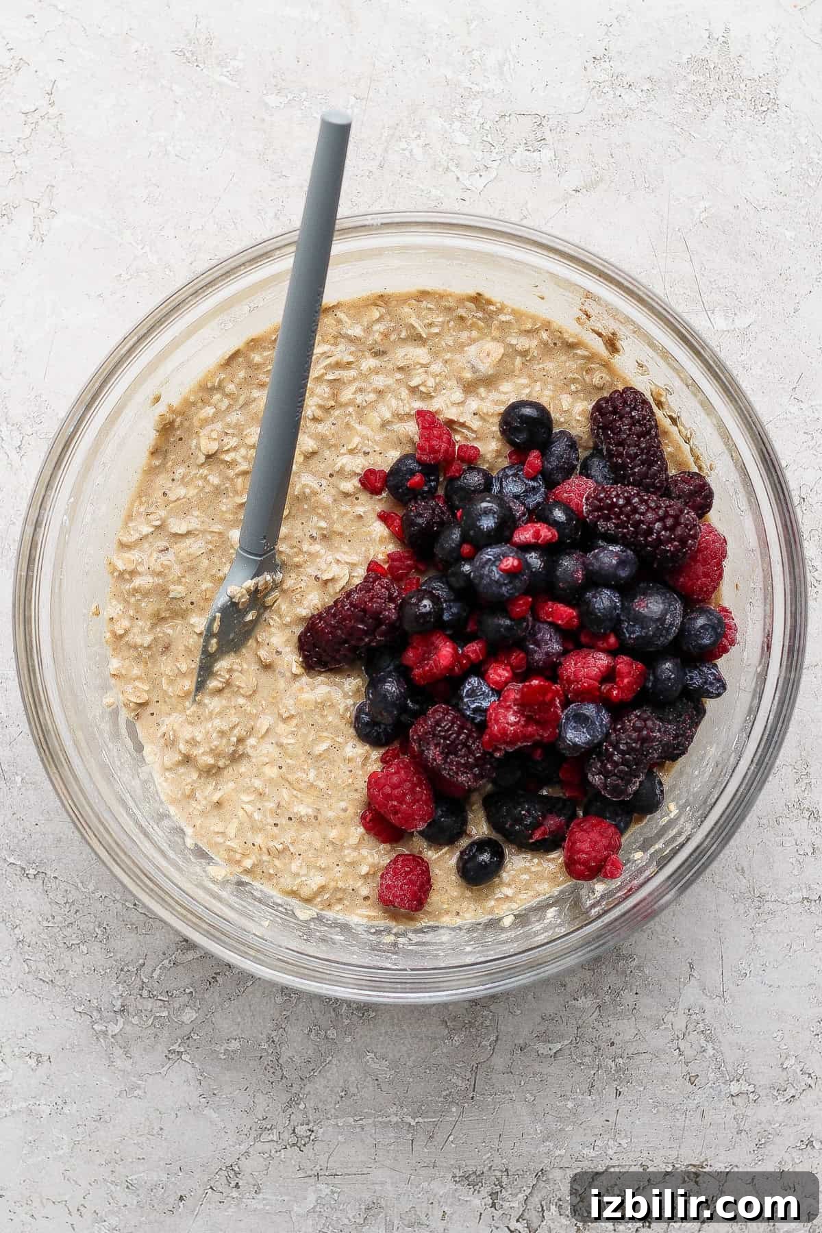 A glass bowl containing oatmeal batter with a pile of mixed berries and a spoon resting inside, placed on a light textured surface.
