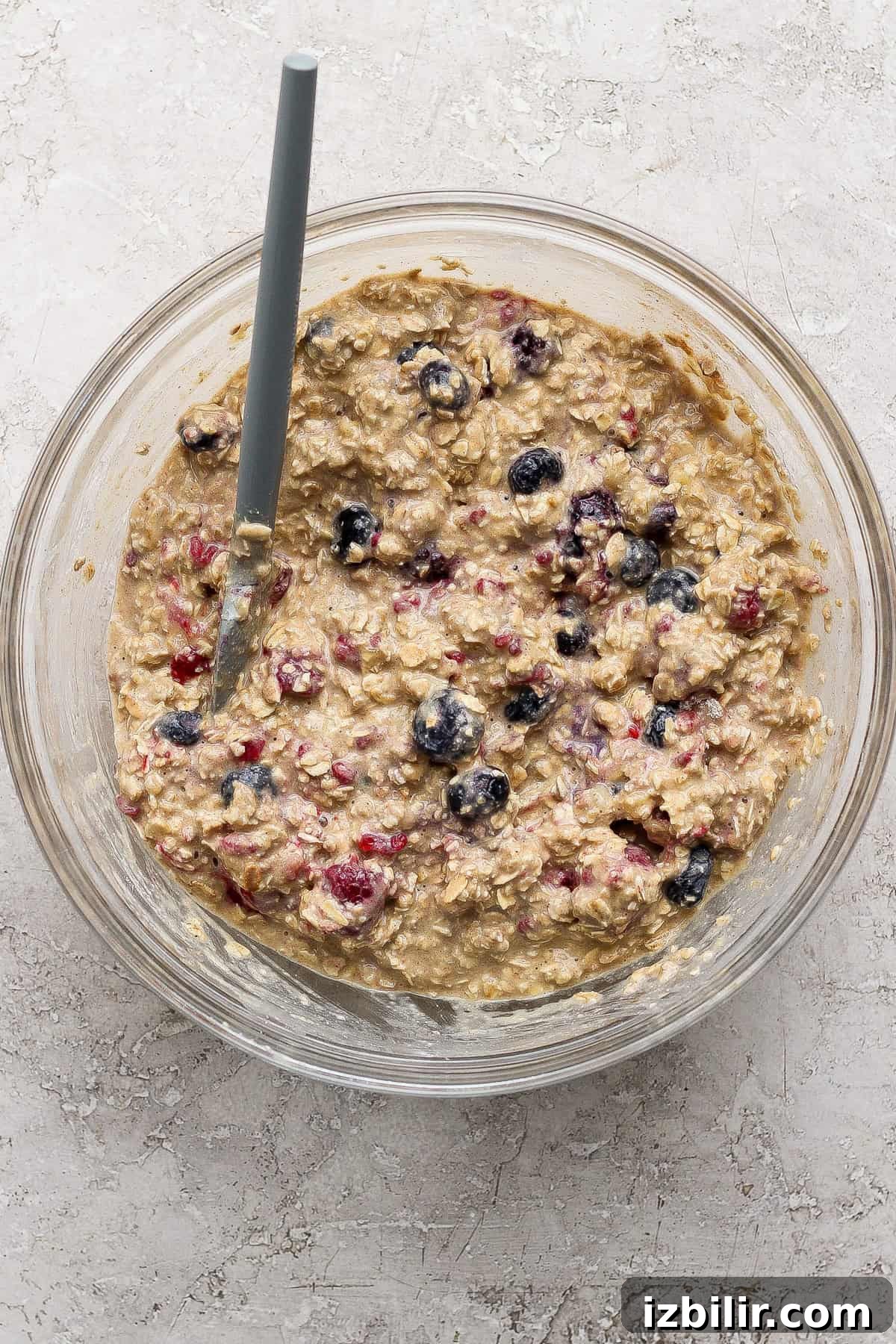 A glass bowl filled with oatmeal batter mixed with berries, with a spoon resting inside, on a light textured surface.