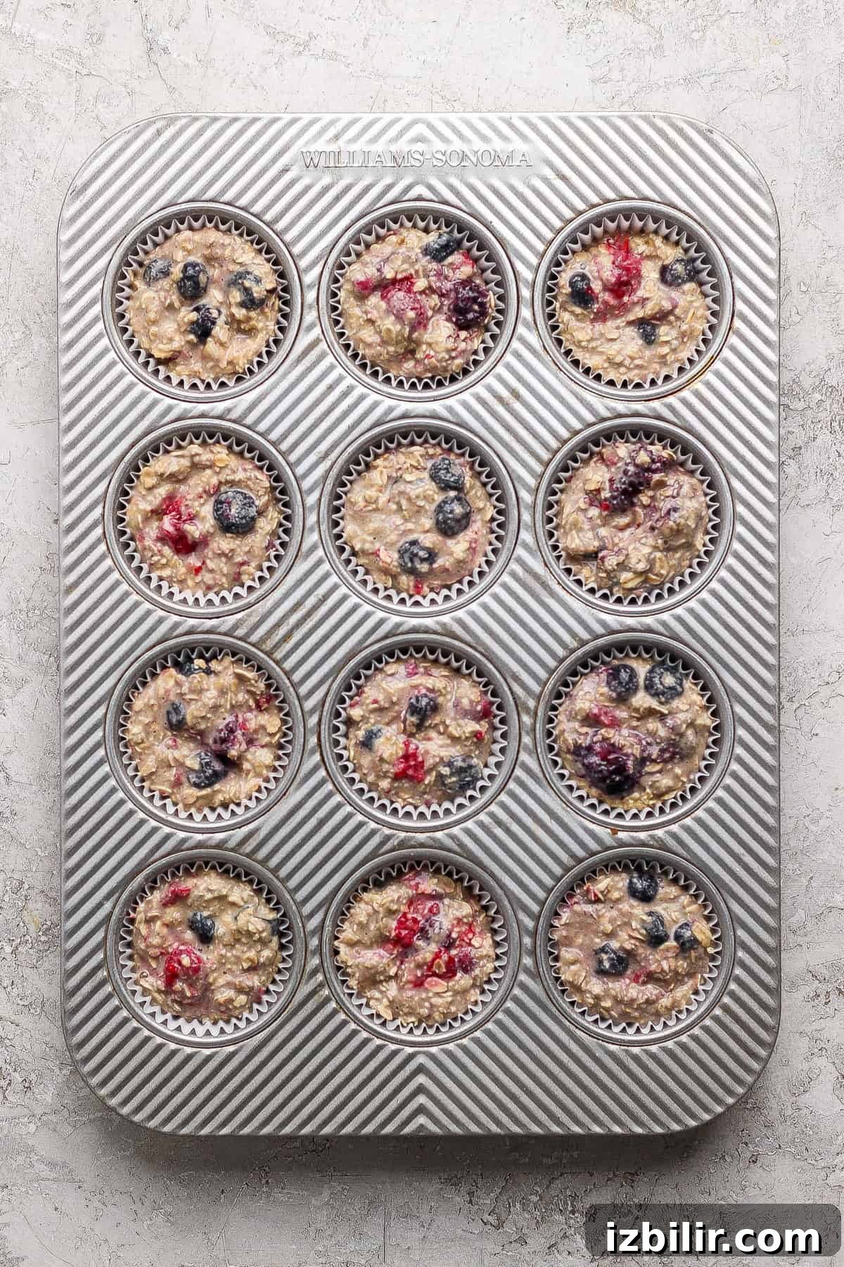 A muffin tin with twelve paper liners filled with unbaked oatmeal batter and mixed berries, placed on a light textured surface.