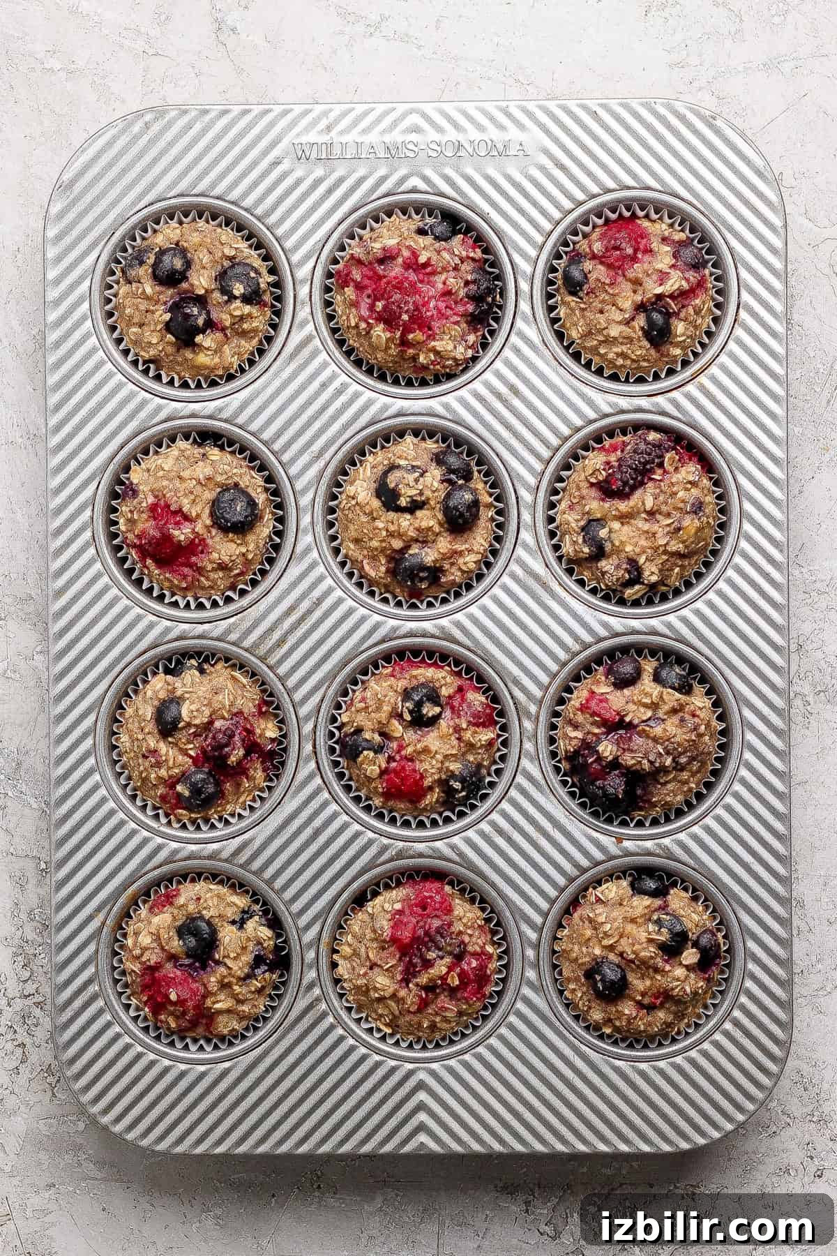 A muffin tin filled with twelve unbaked oat muffins, topped with blueberries and raspberries, on a light-colored surface.
