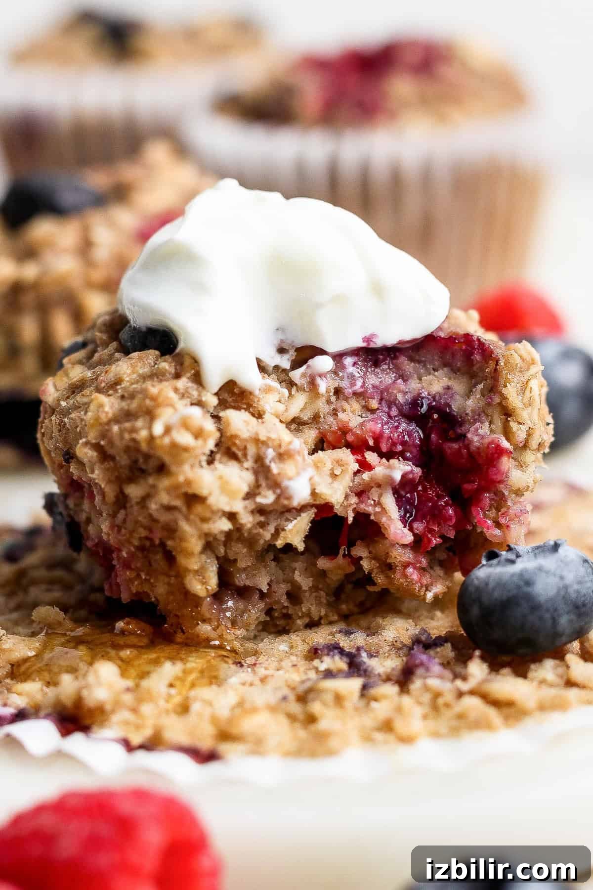 A close-up of a berry muffin with a bite taken out, topped with a dollop of cream, surrounded by fresh blueberries and raspberries.