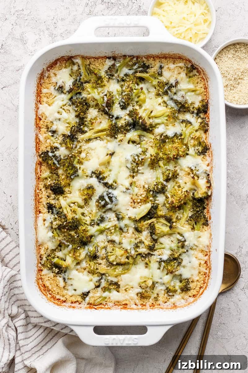 Baked broccoli casserole with melted cheese in a white dish, next to bowls of grated cheese and breadcrumbs on a textured surface.