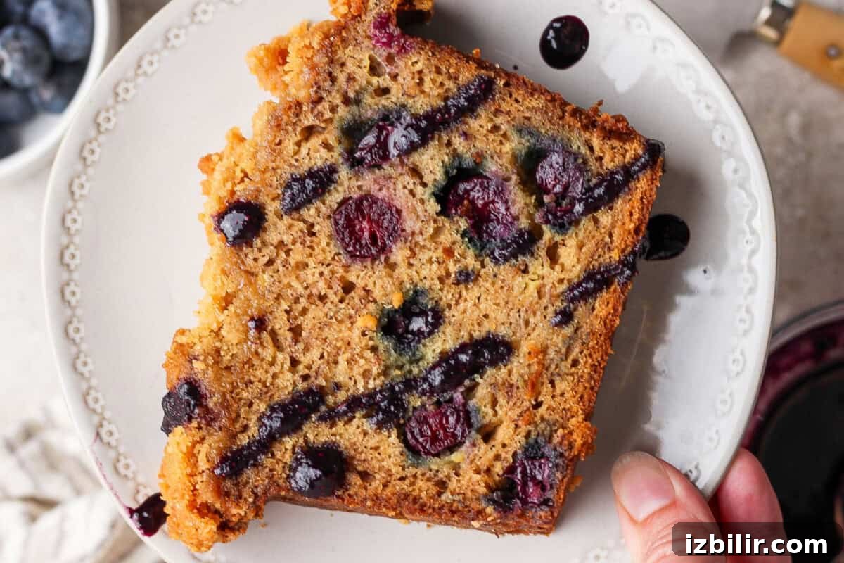 A hand holds a slice of blueberry bread on a white plate, showing blueberries and swirls of blueberry sauce throughout the slice.