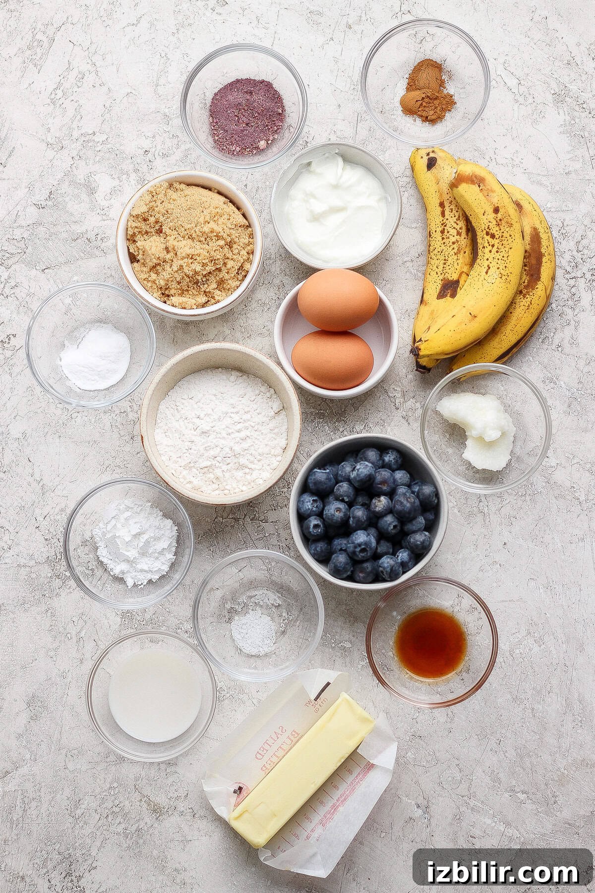 Various baking ingredients in bowls, including flour, brown sugar, eggs, ripe bananas, blueberries, butter, yogurt, vanilla extract, spices, baking powder, baking soda, and coconut oil.