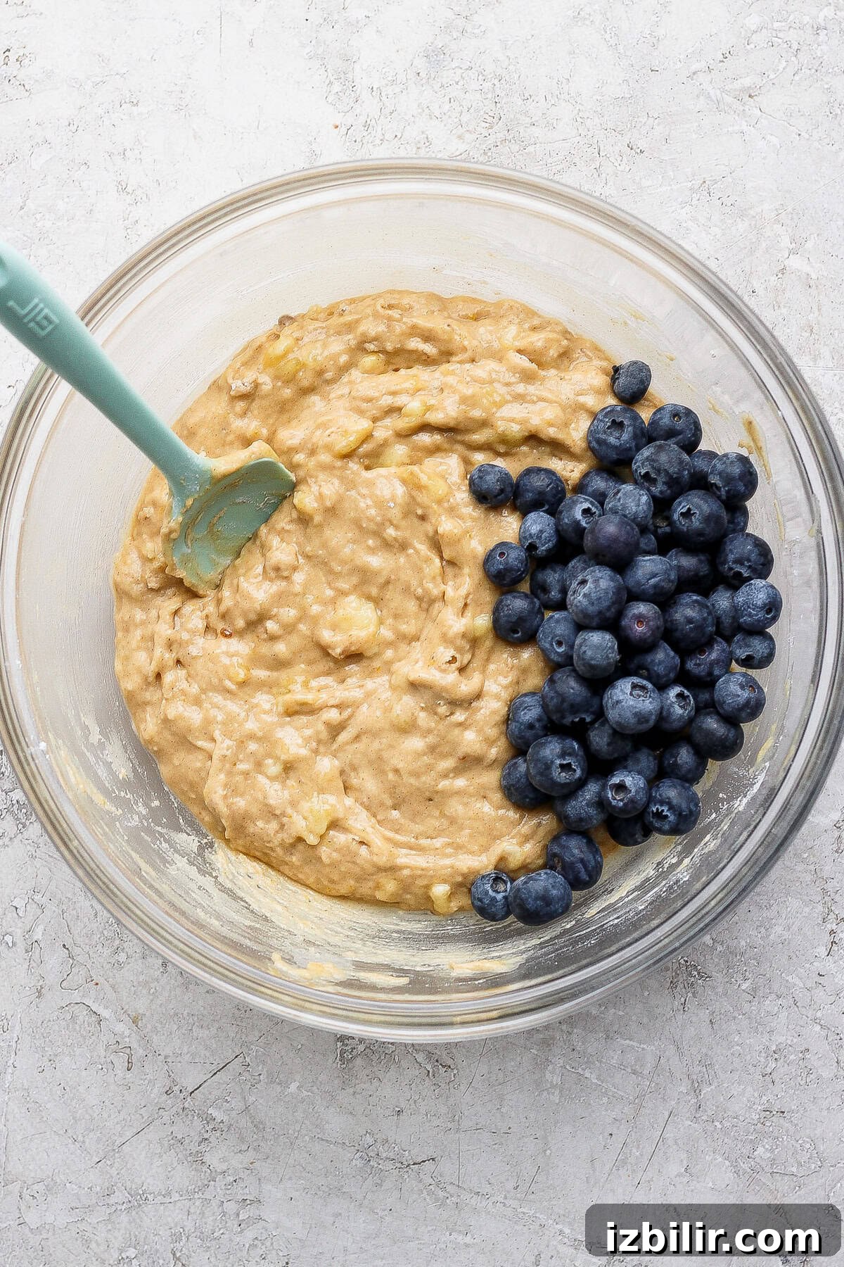 A glass bowl with muffin batter and fresh blueberries, being mixed with a green spatula on a light textured surface.
