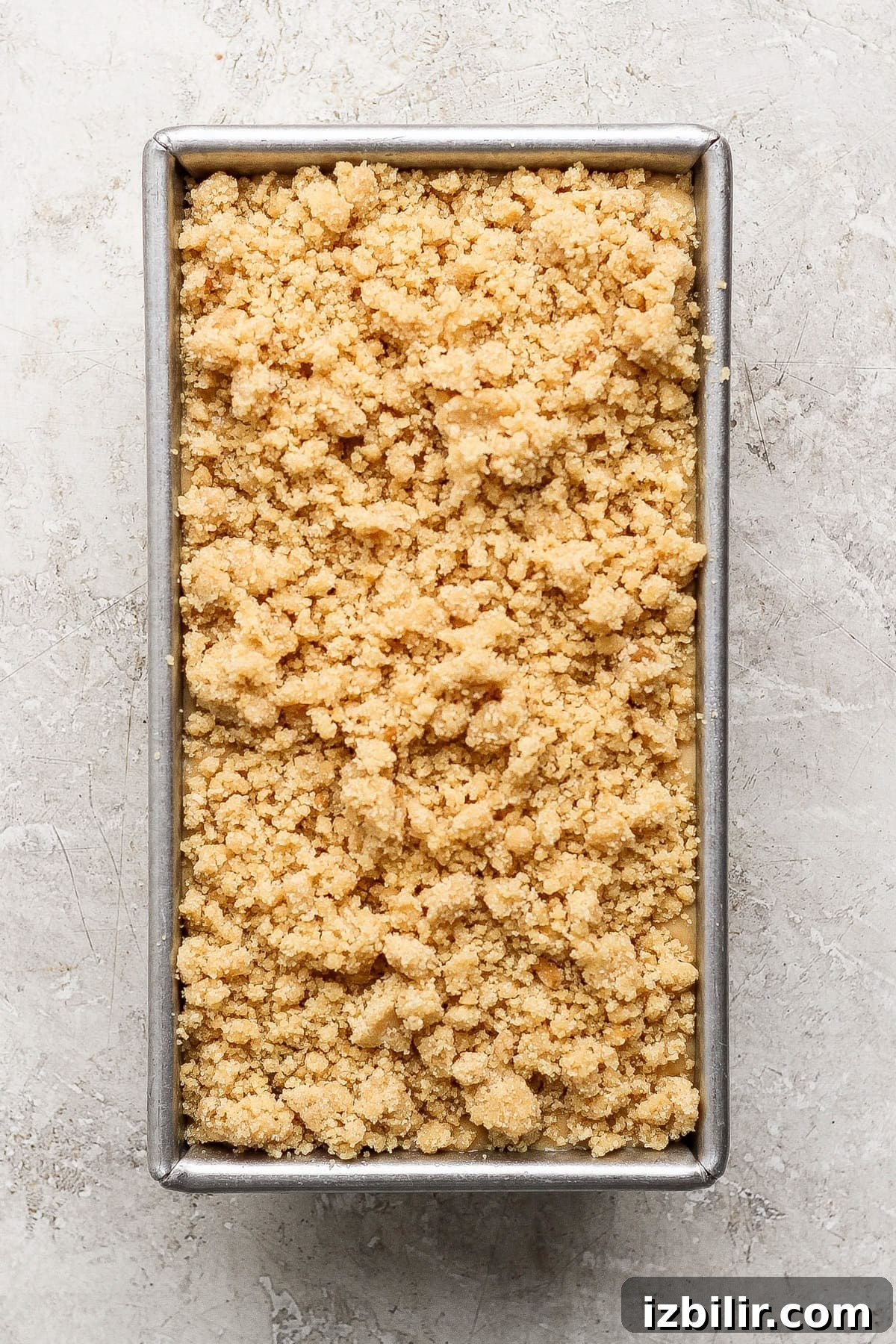 Rectangular metal baking pan filled with an even layer of unbaked, crumbly streusel or crumble topping on a light stone surface.