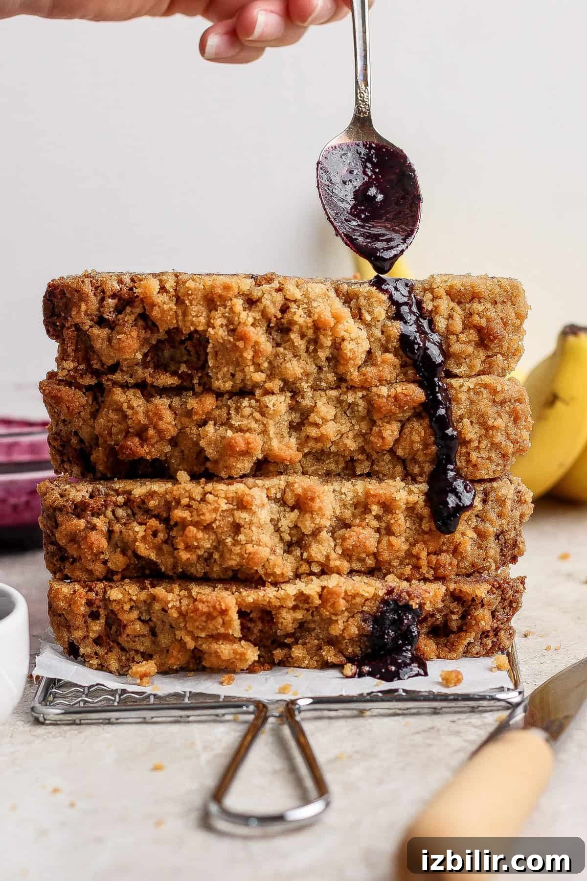 Four slices of crumb-topped bread are stacked on a wire rack as a hand drizzles dark syrup or sauce from a spoon onto the stack. Bananas are visible in the background.