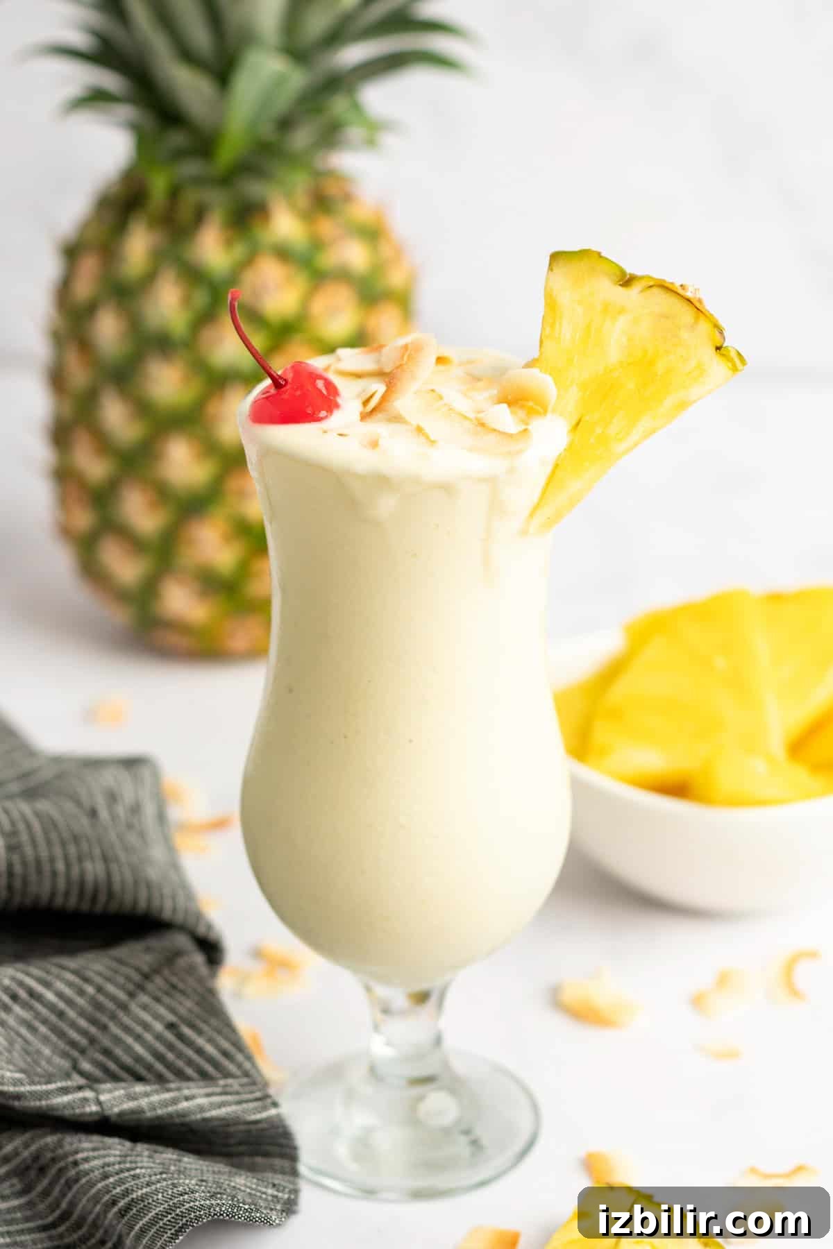 A tall glass of creamy Pina Colada garnished with a pineapple slice, cherry, and shredded coconut, with a whole pineapple and bowl of pineapple chunks in the background.