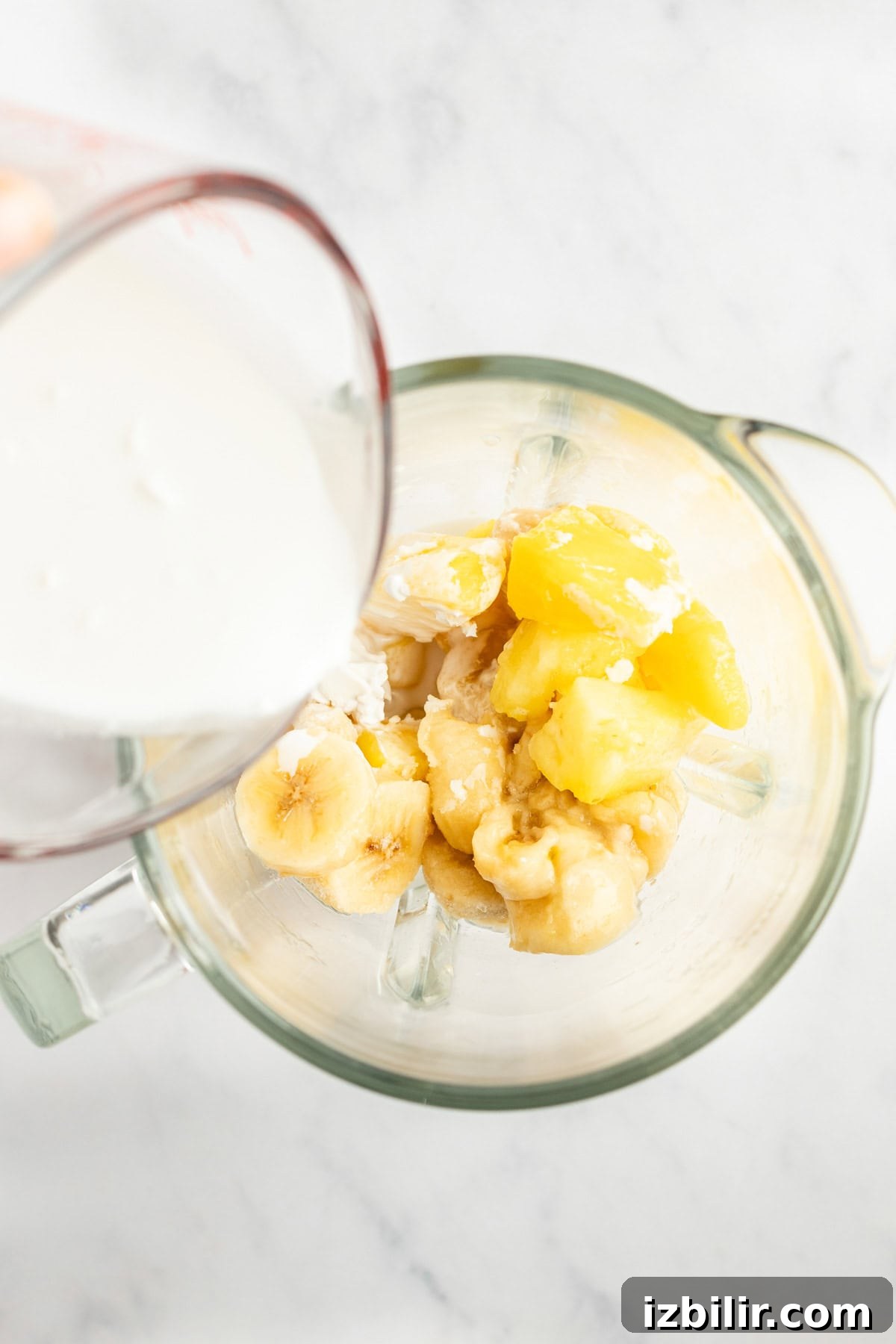A measuring cup pours milk into a blender containing sliced bananas and pineapple chunks.