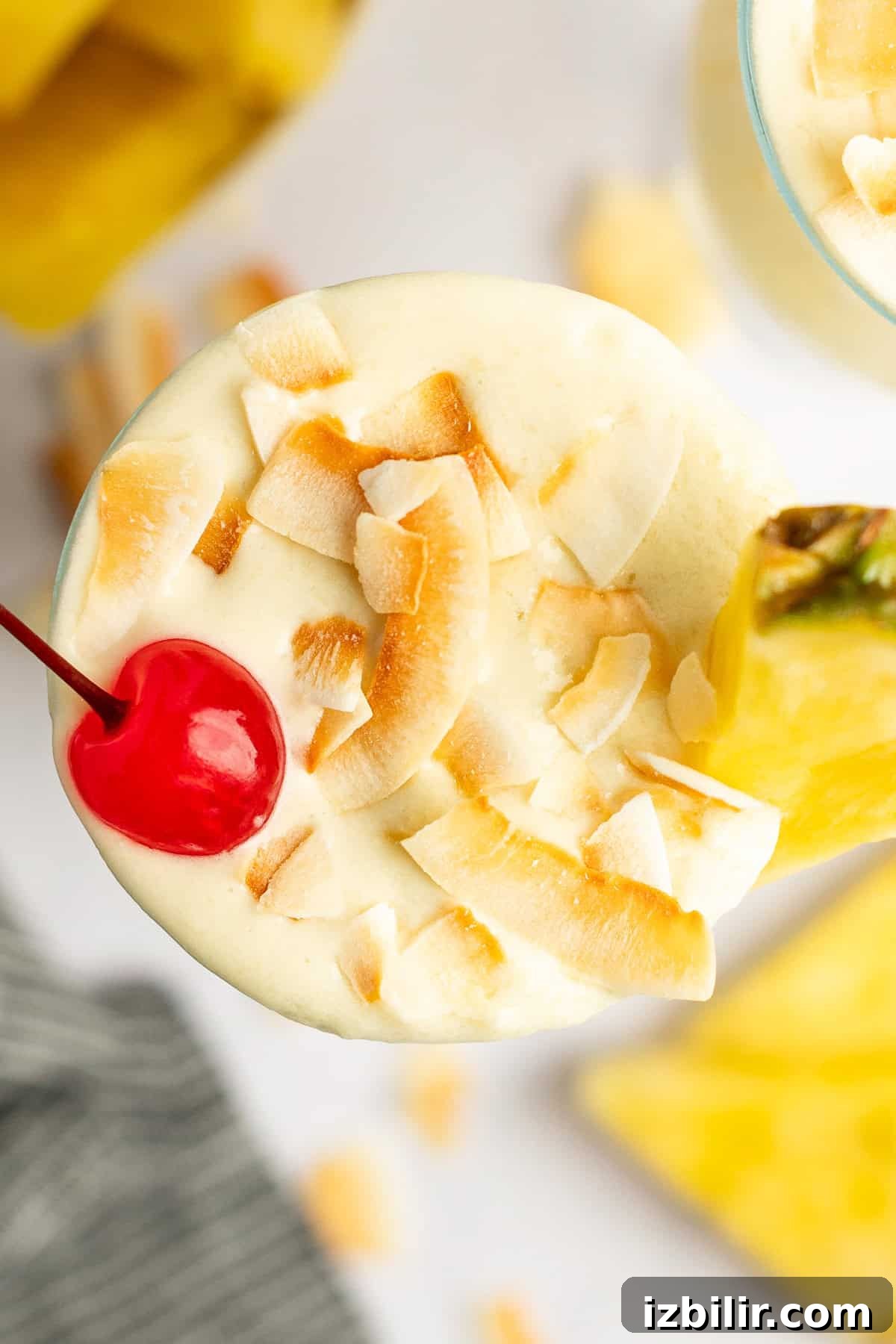 Top view of a creamy cocktail garnished with toasted coconut flakes, a maraschino cherry, and a pineapple wedge.