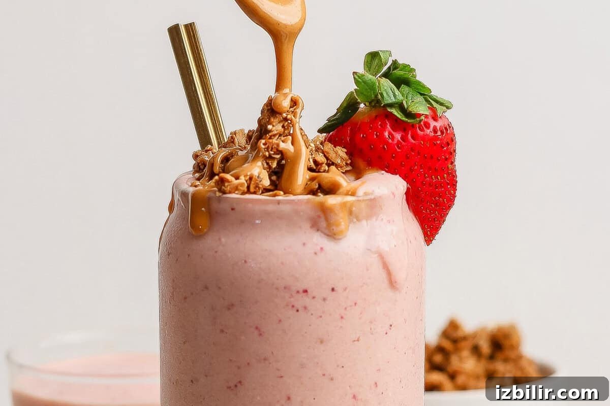 A strawberry smoothie topped with granola, peanut butter, a fresh strawberry, and two straws in a glass.